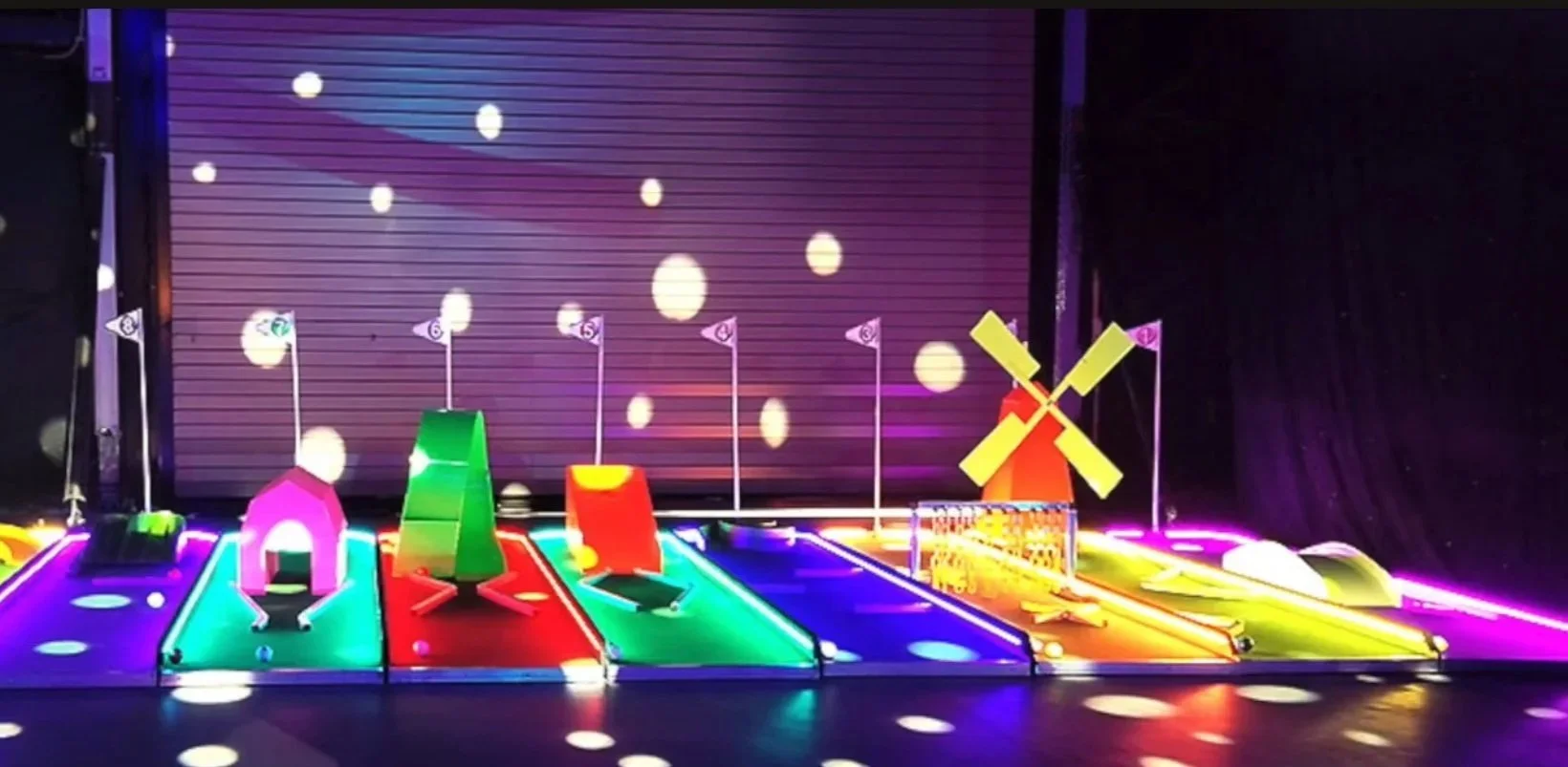 Miniature colorful golf course with small windmill, flags, and play structures illuminated with bright neon lights.
