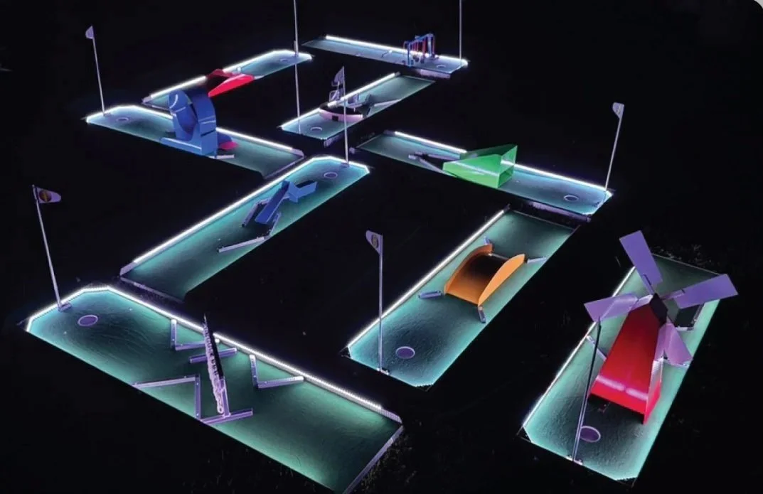 Miniature golf course with illuminated pathways, colorful windmill attractions, and flags on each hole set against a dark background.