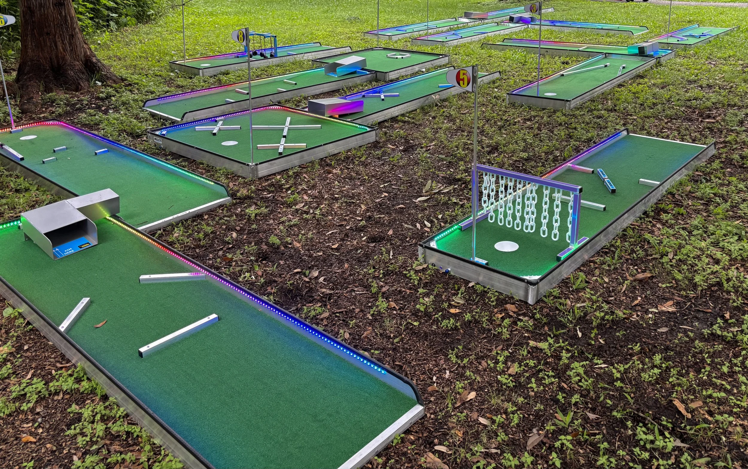 Miniature golf course with various holes, obstacles, and LED lights, set outdoors on grass and dirt ground.