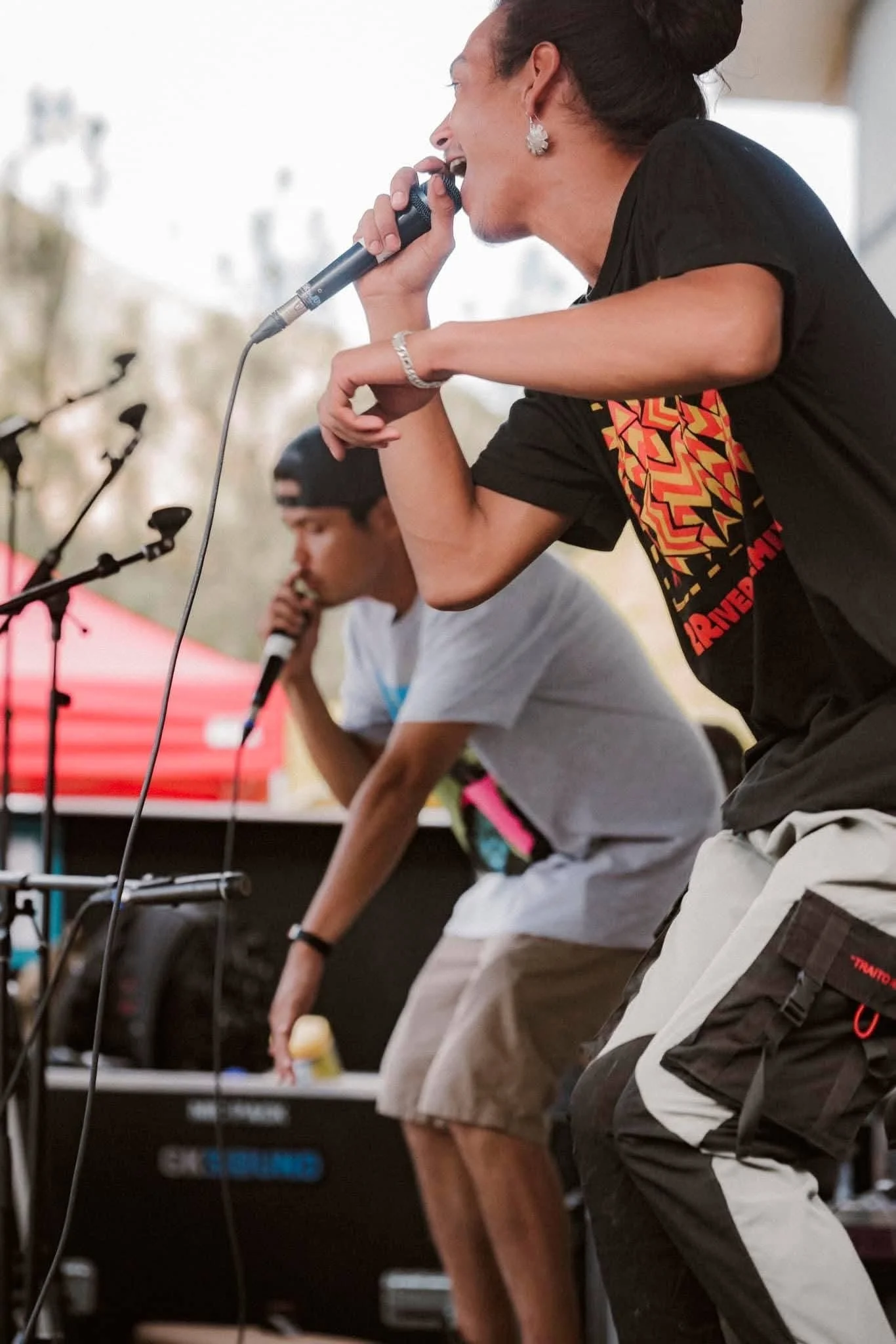 A performer singing into a microphone on stage, with another musician in the background playing an instrument. Both are casually dressed and appear to be engaged in a live performance.
