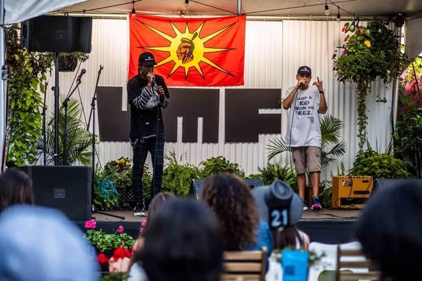 Two performers on an outdoor stage, one singing into a microphone and the other gesturing with hand, with a red and yellow banner featuring a fist and sunburst behind them. Audience seated in front, some wearing hats and glasses, watching the perform