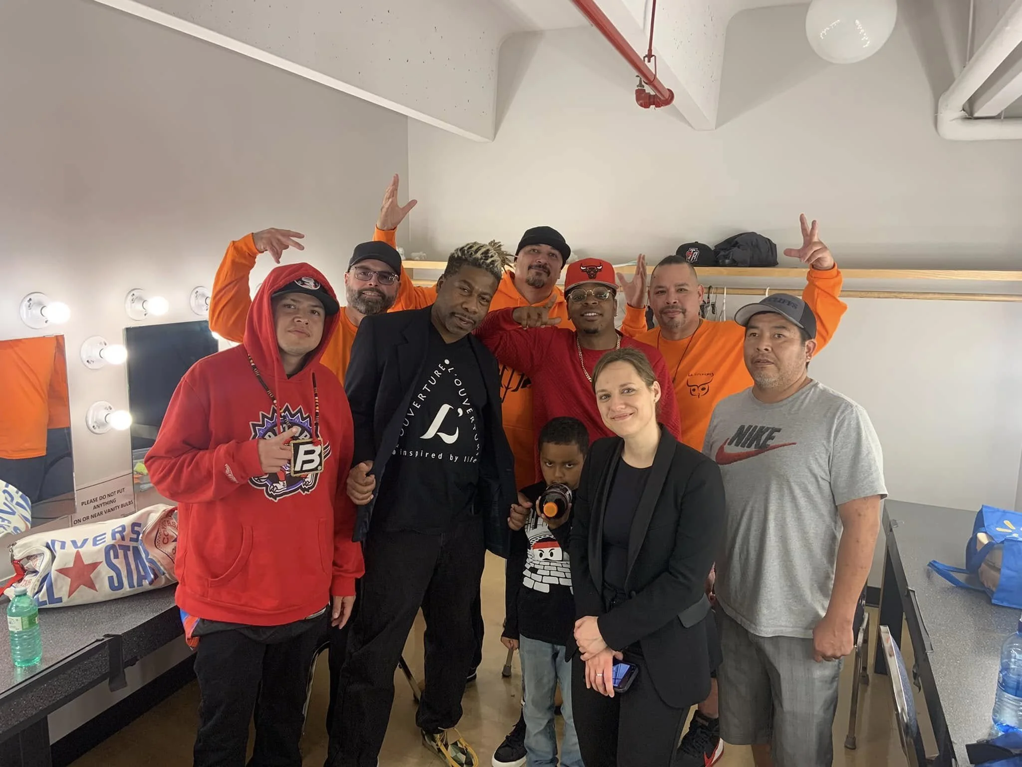 Group of men and women posing together indoors, some making peace signs and others smiling, behind them are mirrors, lights, and various bags and items on a counter.