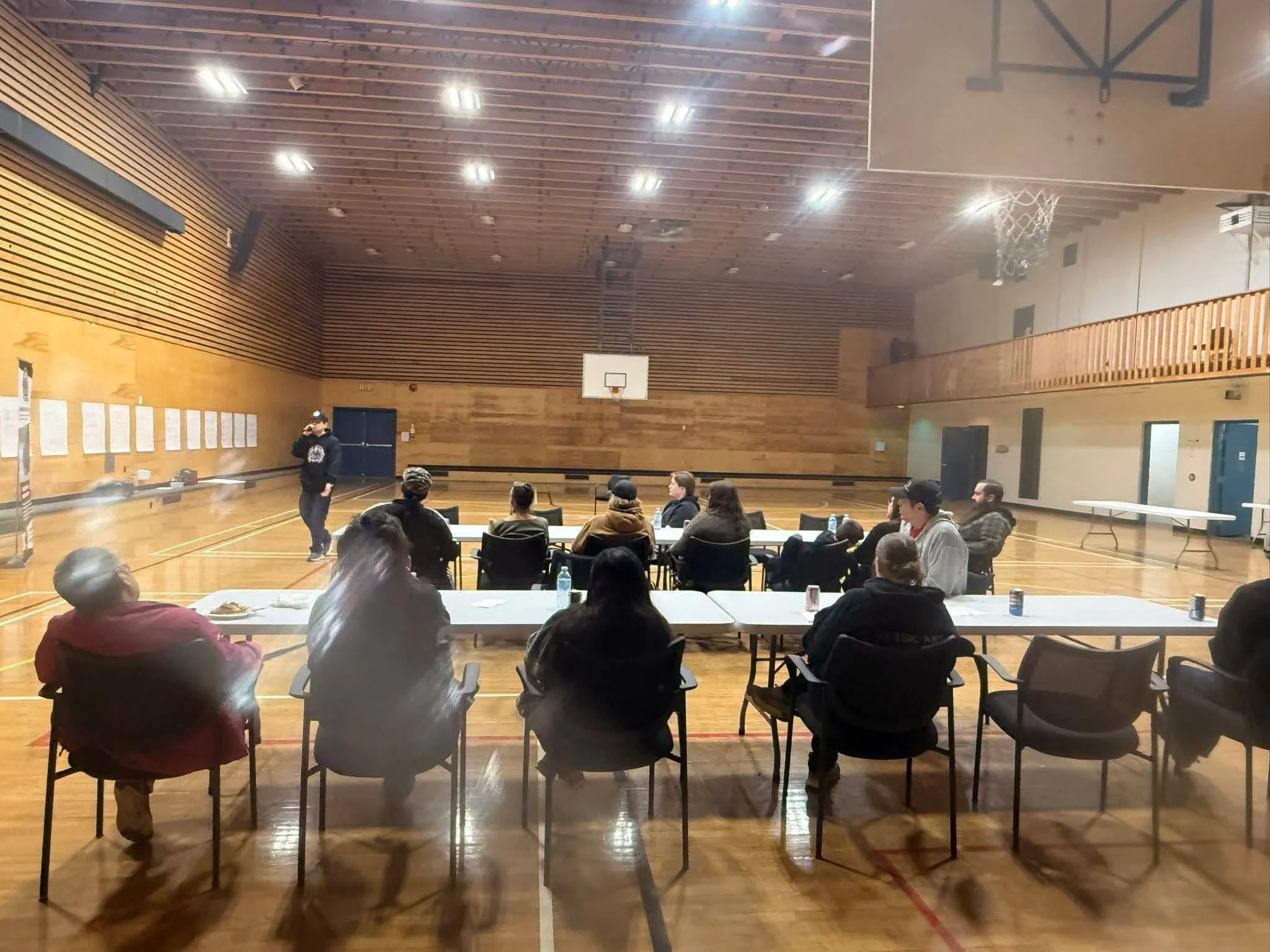 People seated at tables in a gymnasium, with a person speaking at the front, wooden walls, and a basketball hoop.