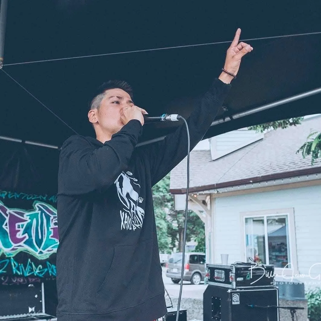 A young man singing into a microphone on an outdoor stage, with his right arm raised and finger pointing upward. He is wearing a black hoodie with a graphic design, and there are speakers and a backdrop behind him. A house and parked cars are visible
