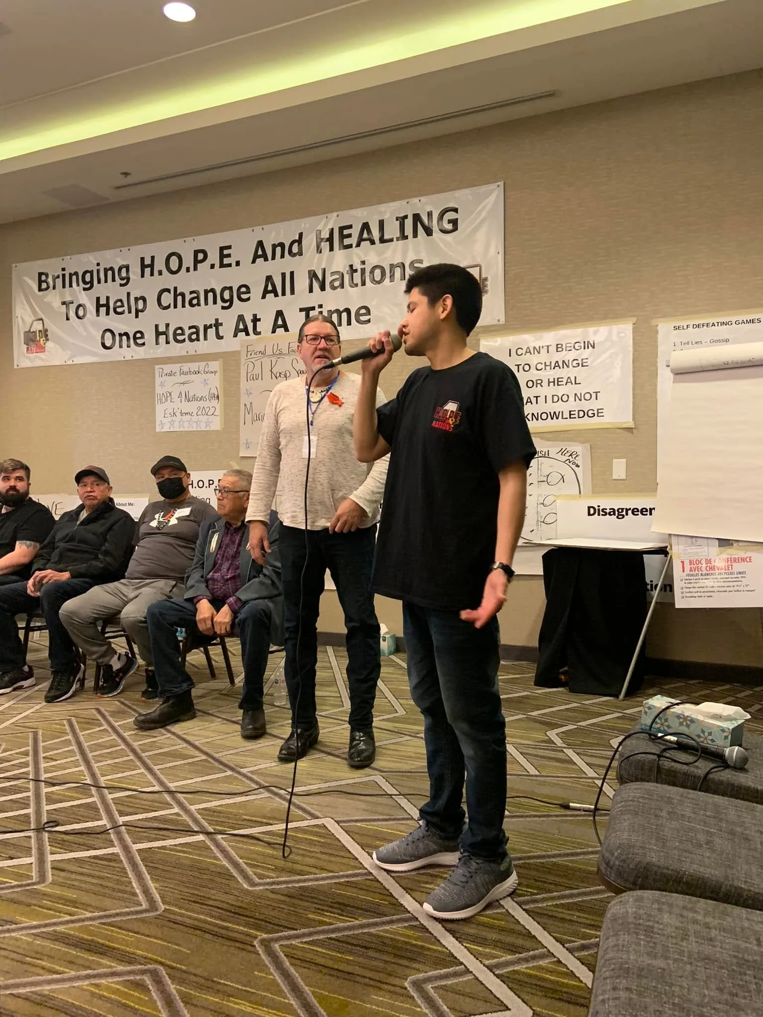 A young man speaking into a microphone at a conference event, standing in front of a seated audience. There are banners and posters on the wall with messages about hope, healing, and change. The room has a patterned carpet and chairs arranged in rows