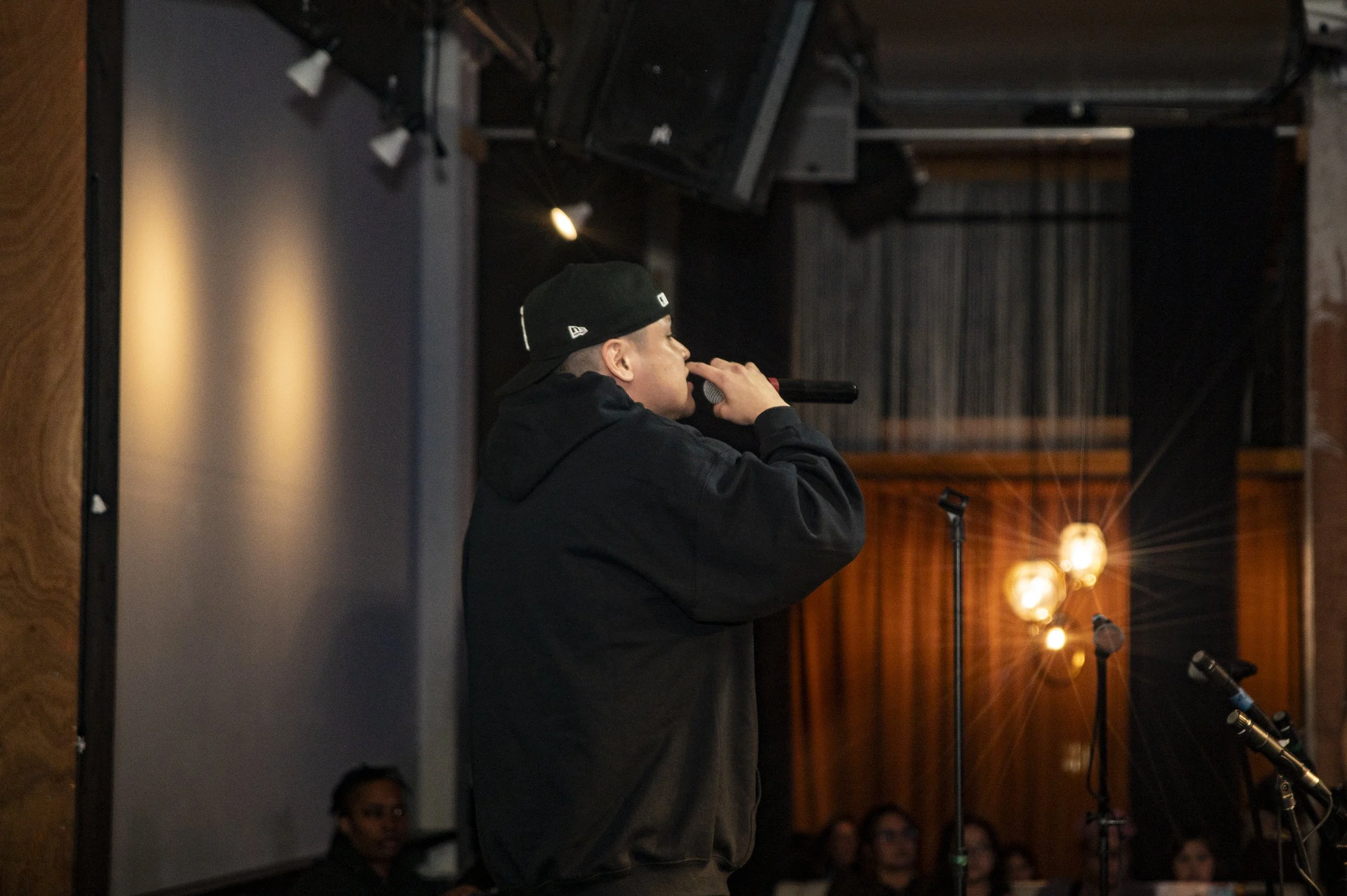 A person wearing a black cap and jacket is holding a microphone and appears to be performing or speaking during an event. The setting seems to be indoors, with warm lighting and several people visible in the background.