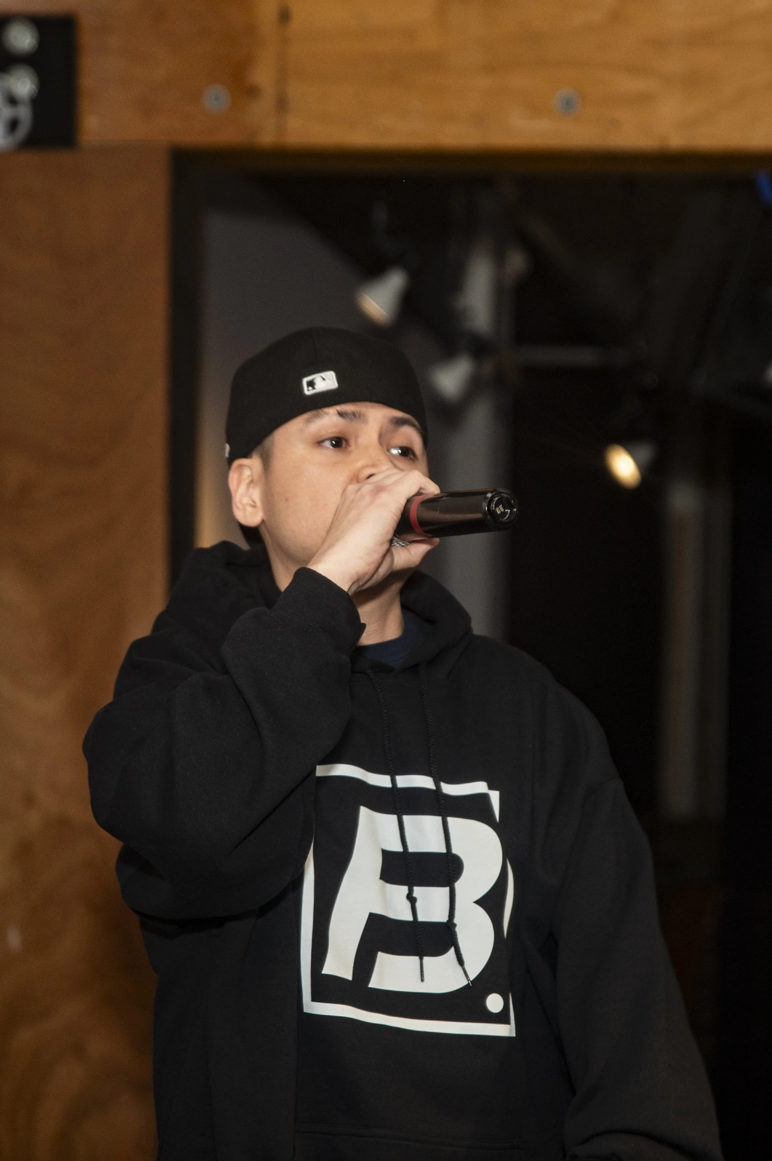 A young man wearing a black hoodie with a white graphic design and a black beanie, holding a microphone and standing indoors, with wooden wall panels in the background.