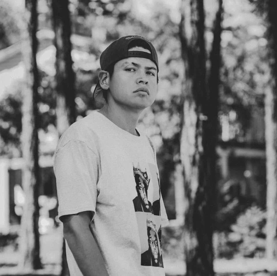 Young man with a serious expression, wearing a backward cap and a graphic T-shirt, standing outdoors in front of trees.