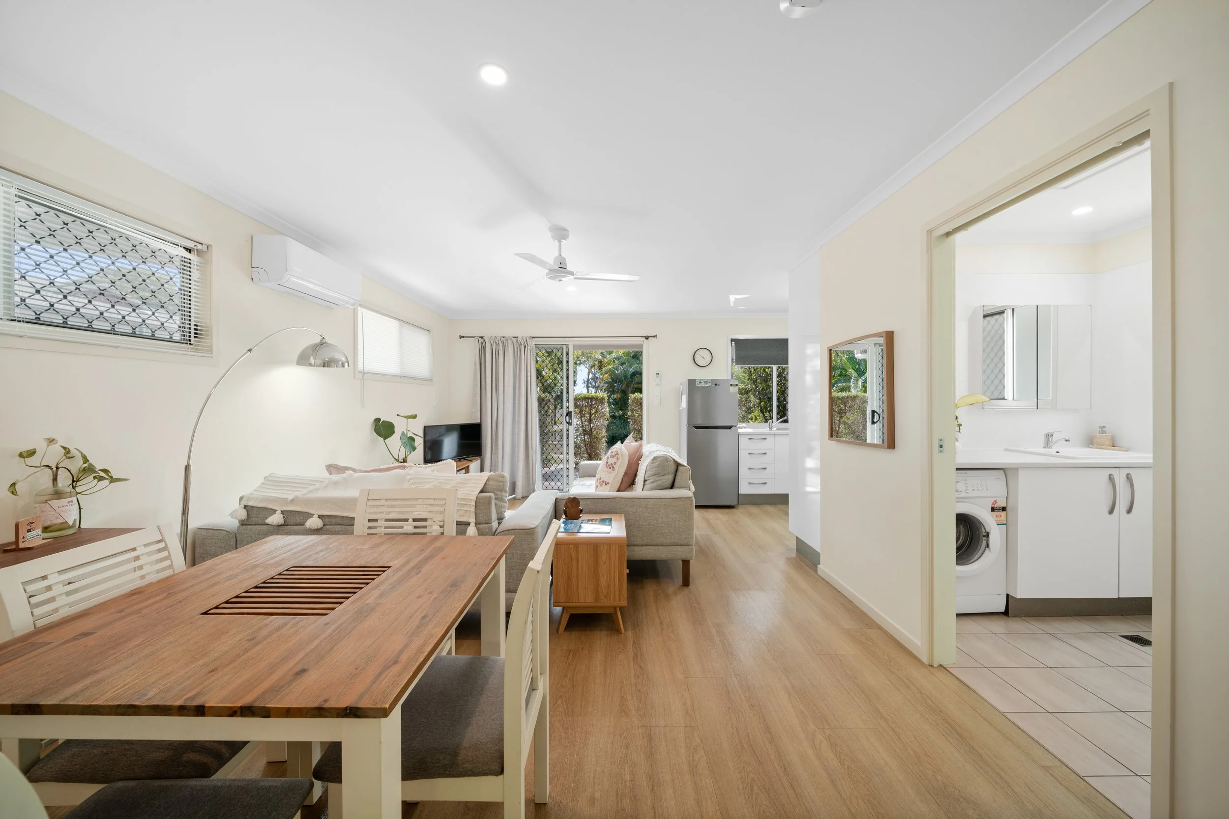 Interior of a living room with a dining table in the foreground, beige walls, large windows, a ceiling fan, a small TV, a sofa, plants, and an open kitchen area with a washing machine.