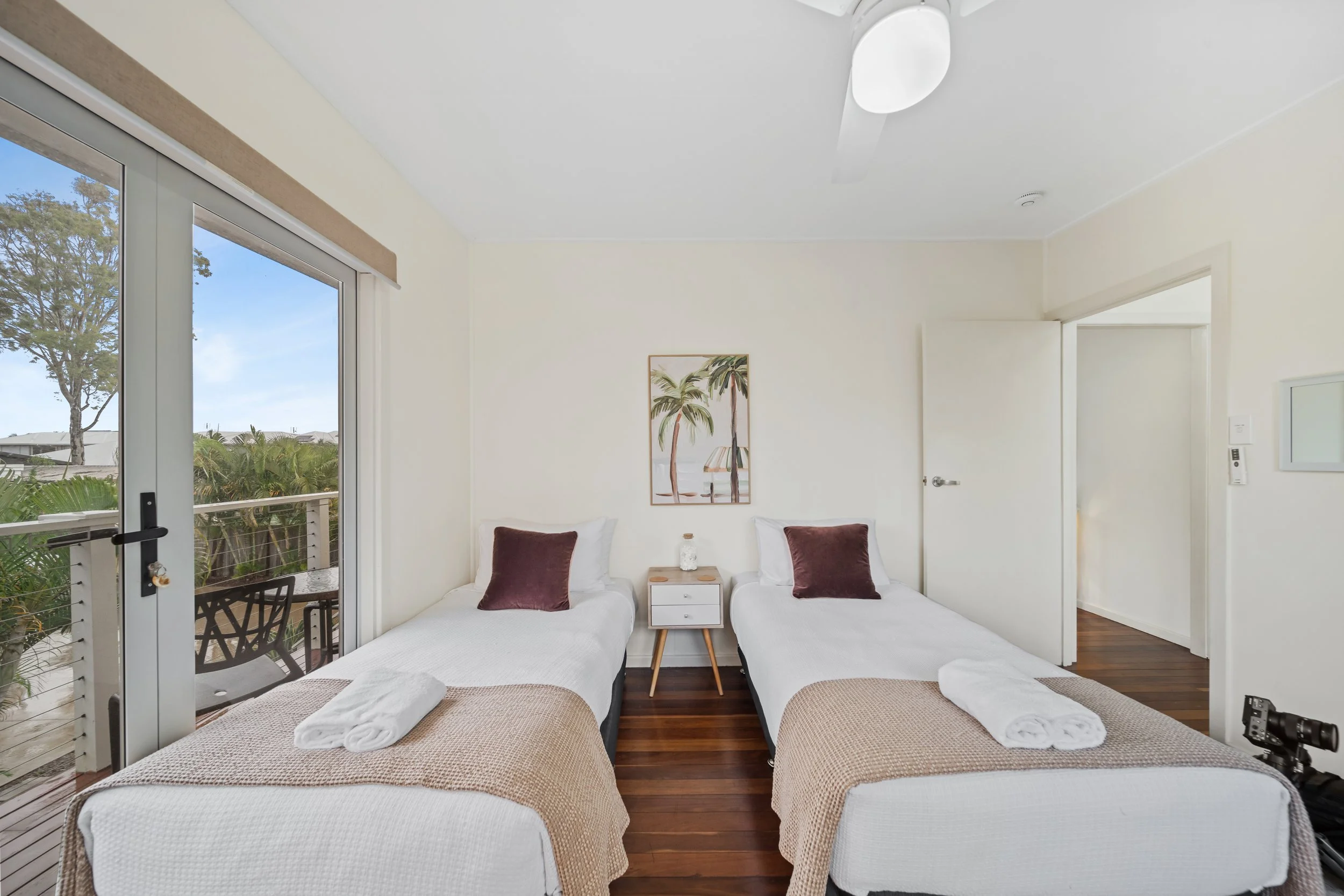 A room with four beds, two on each side, with towels and pillows on top. A small white nightstand is between the beds, with a picture of palm trees hanging on the wall above. There is a sliding glass door leading to a balcony with outdoor furniture a