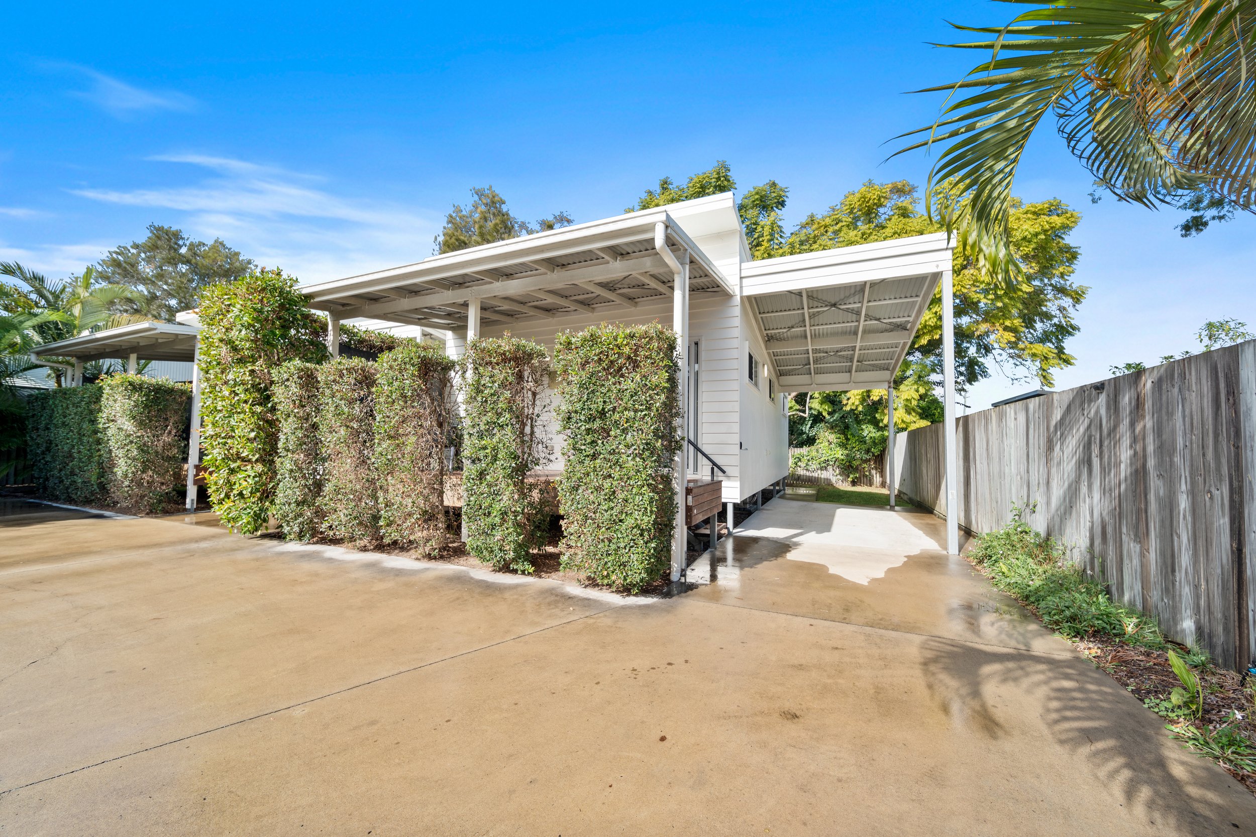 A modern white house with a metal roof, surrounded by lush green trees and bushes, with a concrete driveway and a wooden fence on the right side under a clear blue sky.