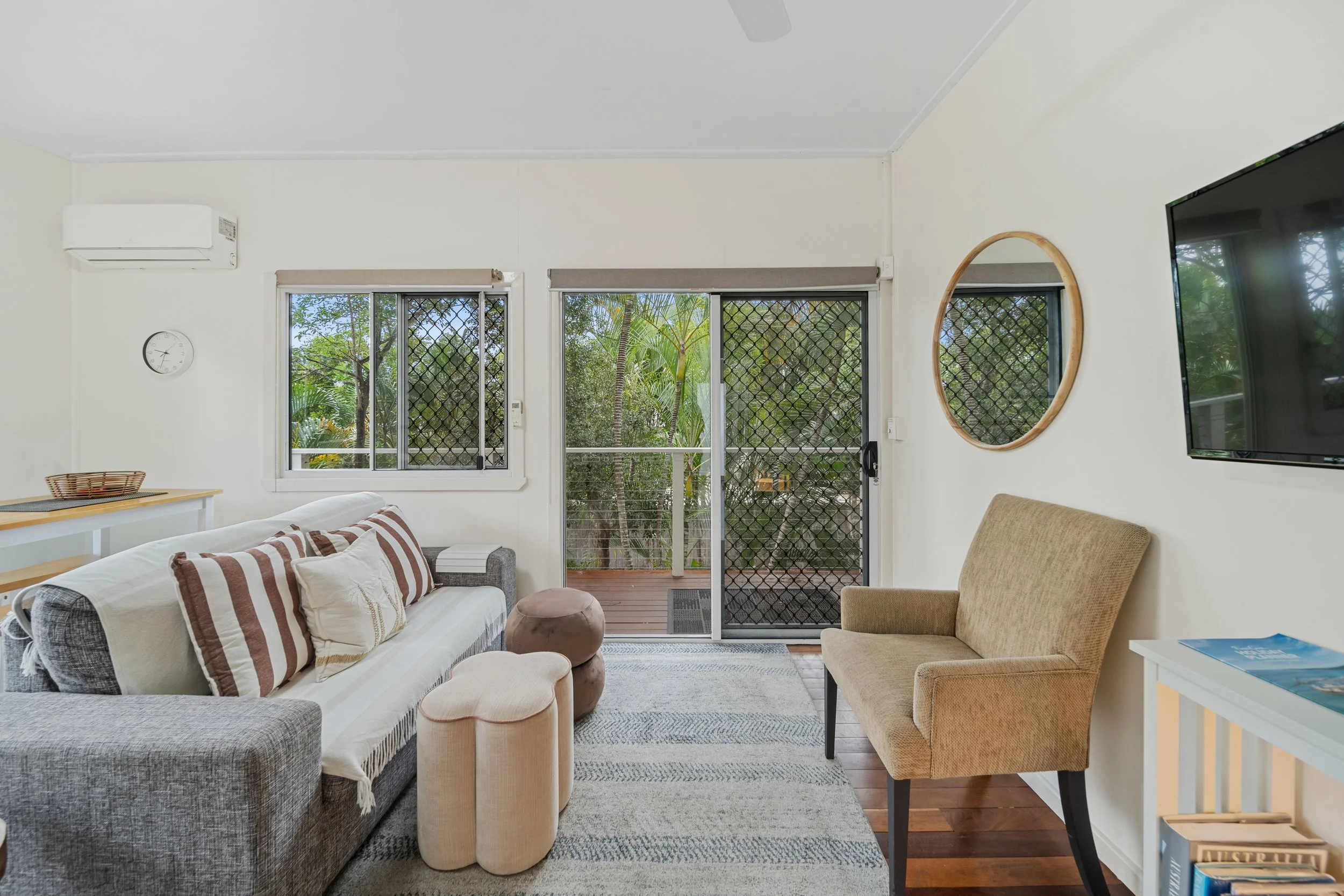 Living room with gray sofa, beige armchair, striped pillows, round ottomans, wall mirror, wall clock, air conditioner, and sliding glass door leading to outdoor patio with greenery.