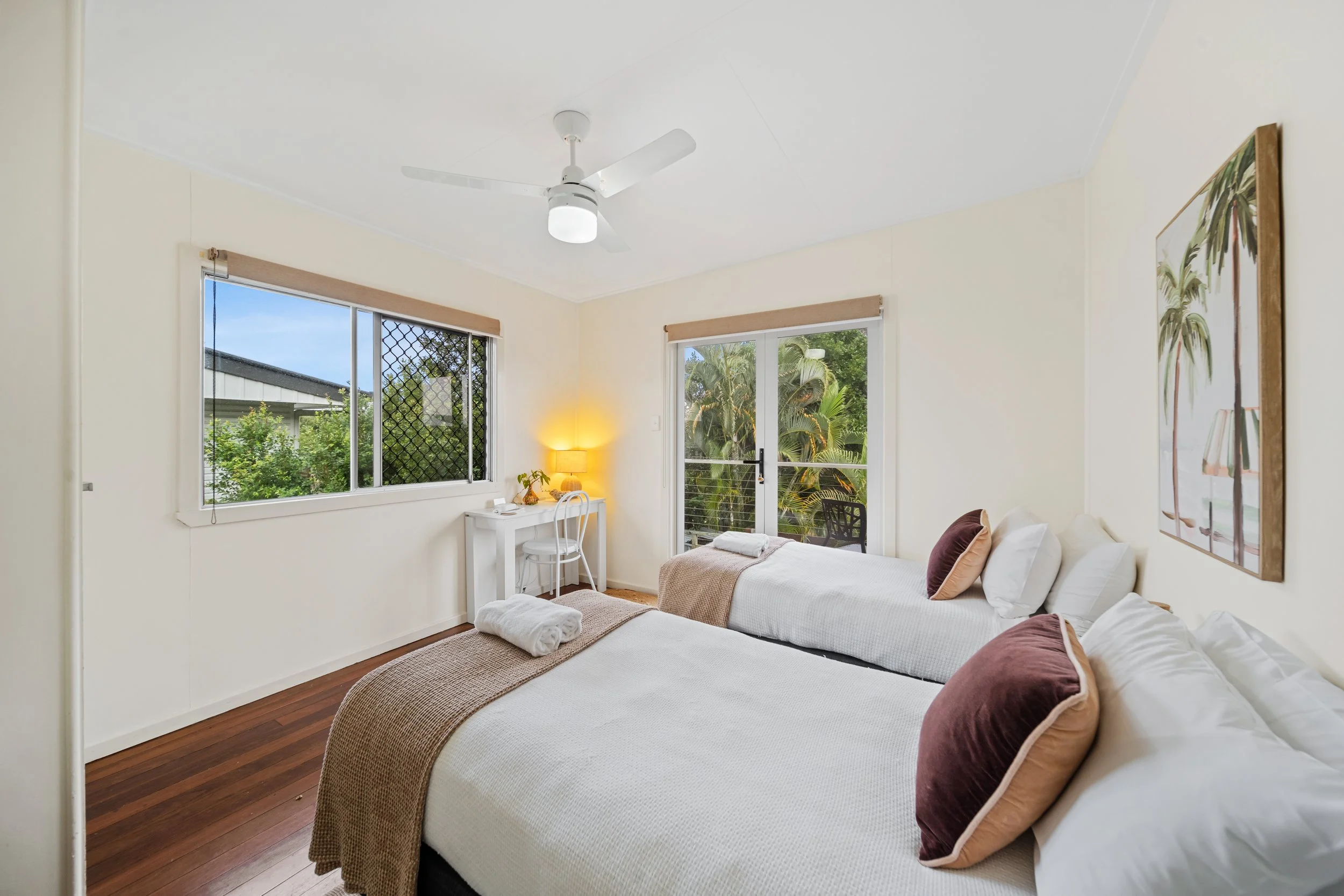 A bright bedroom with two twin beds, white bedding, and brown and white pillows. A window with a view of greenery and a sliding glass door lead to a balcony. There's a small white desk with a white chair, a lamp, and some decorations. A ceiling fan a