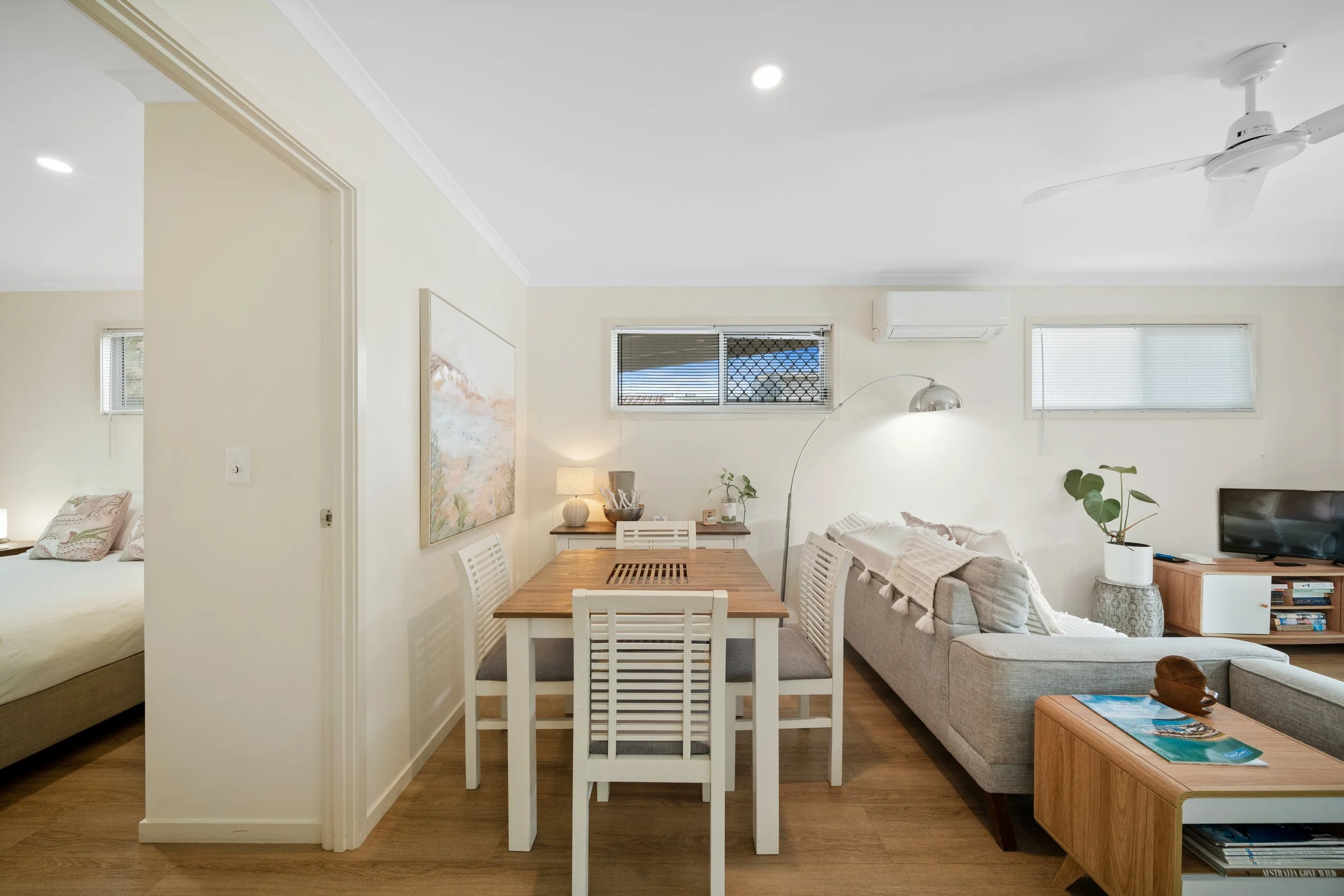 A living room with a white sofa, a wooden coffee table, a dining table with white chairs, a potted plant, and windows with blinds. There's a ceiling fan and an air conditioner on the wall.