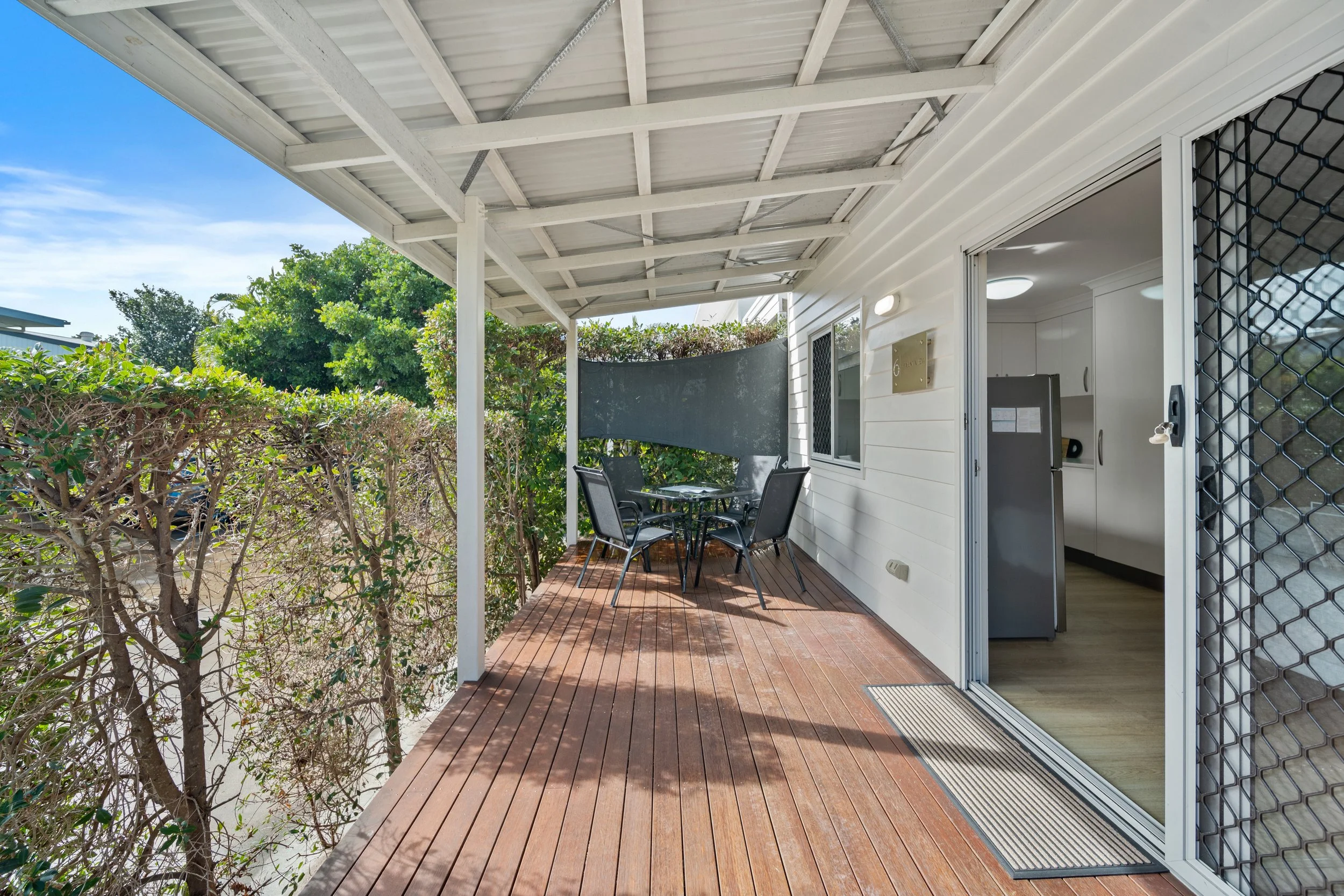 Outdoor patio with wooden flooring, a small round table with chairs, and sliding glass door leading inside a house. There are bushes and trees in the background and a covered roof overhead.
