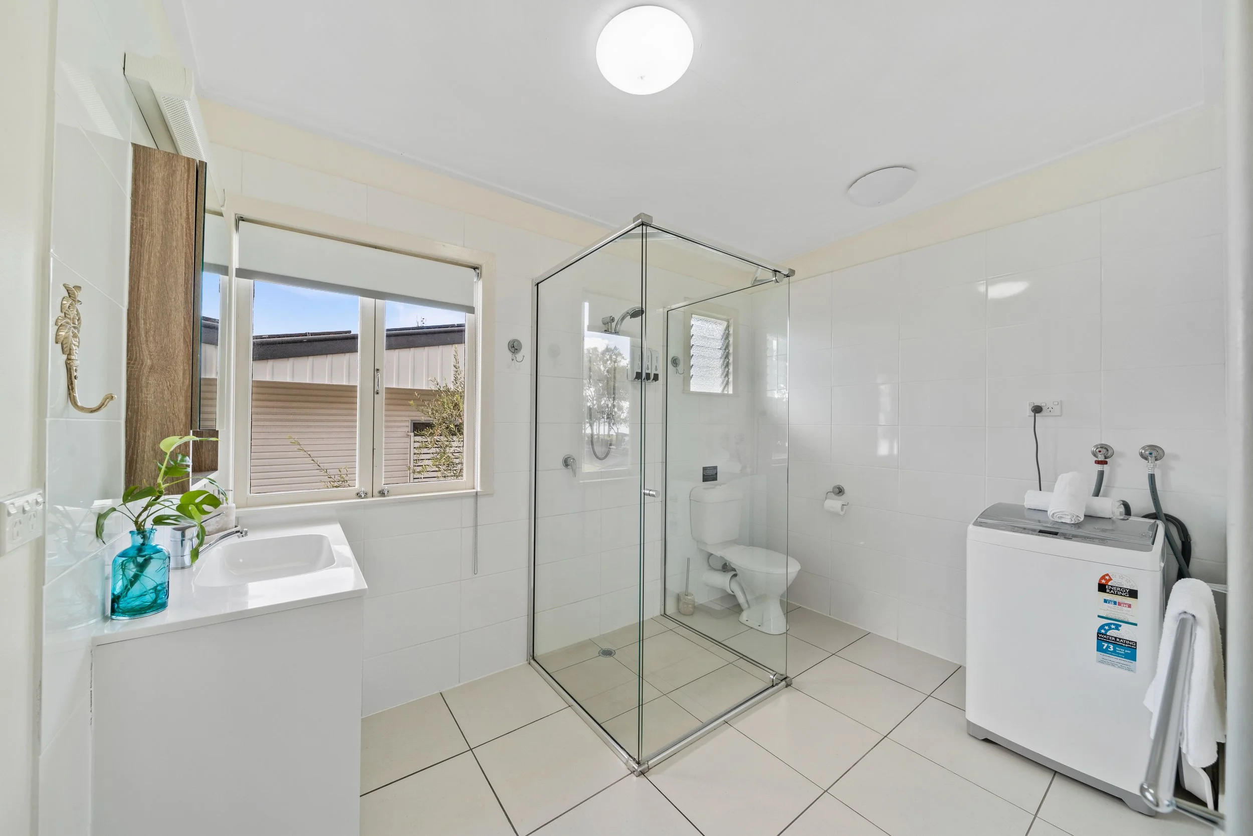 A clean, modern bathroom with a large window. It features a glass-enclosed shower, a white toilet, a washing machine, and a sink with a mirror and decorative plants.