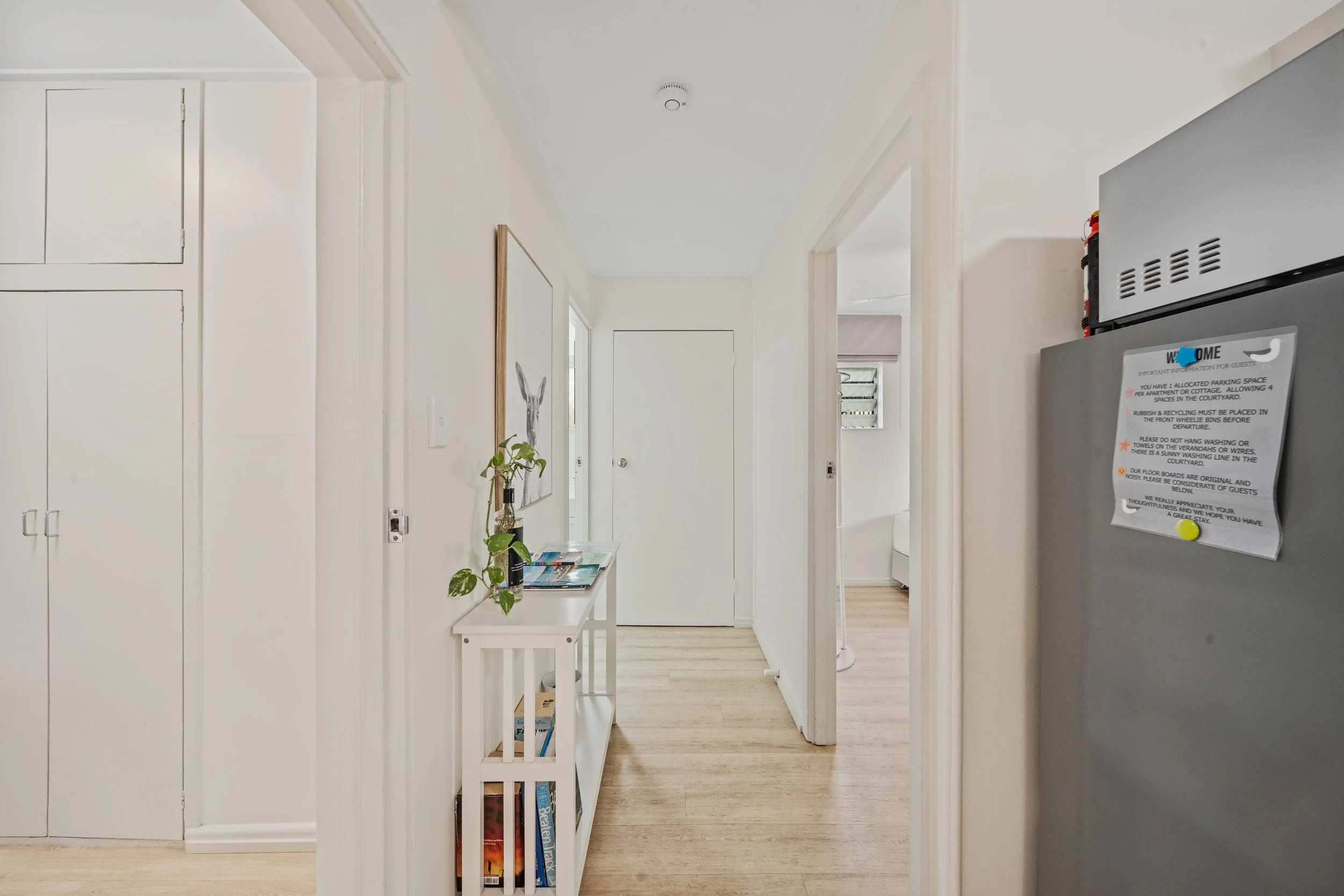 A bright hallway in a modern home with white walls, light wood flooring, a small white table with magazines, a framed artwork of a rabbit on the wall, and a partial view of a stainless steel refrigerator with a whiteboard and magnets.