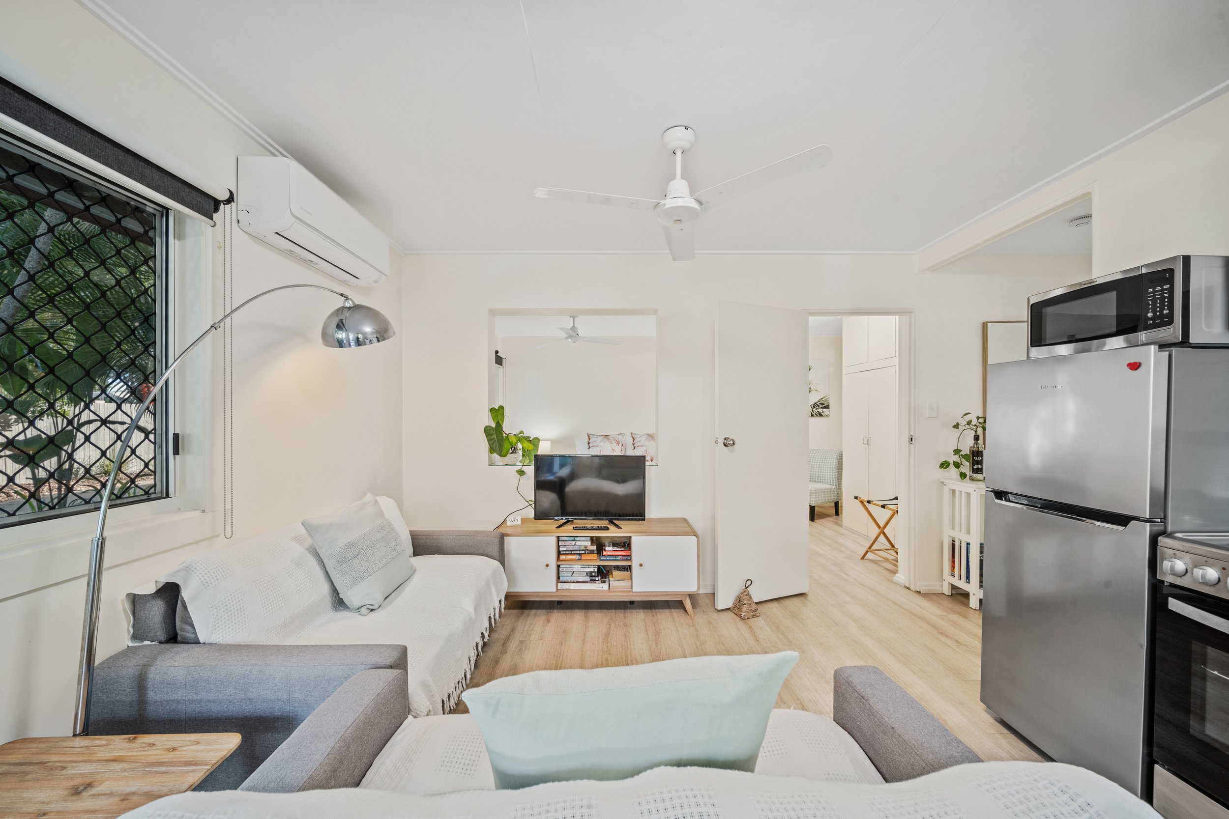Living room with gray sofas, a small TV on a stand, and a white ceiling fan, with a window with security bars, an air conditioner, and a kitchen area with a microwave and refrigerator.