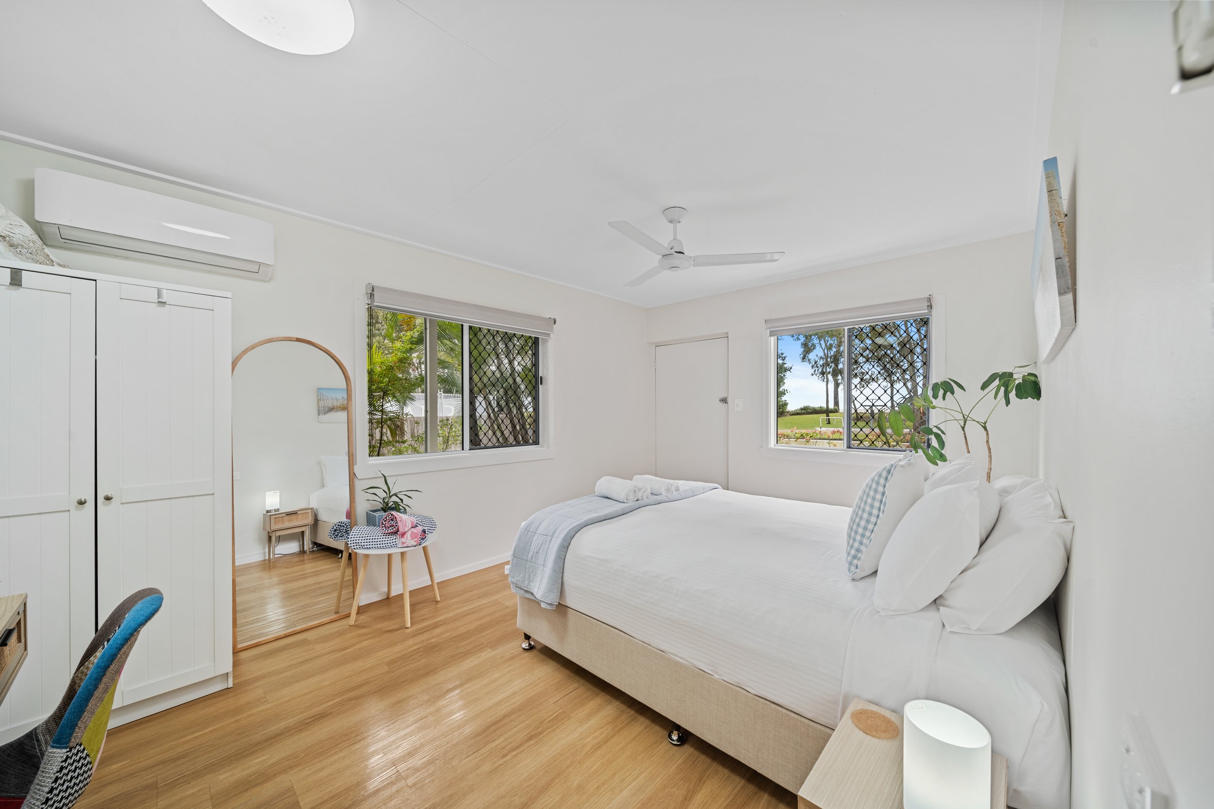 A bright bedroom with white walls, a ceiling fan, windows with security screens, a bed with white linens and pillows, a wooden side table, a potted plant, and a wall-mounted air conditioner.