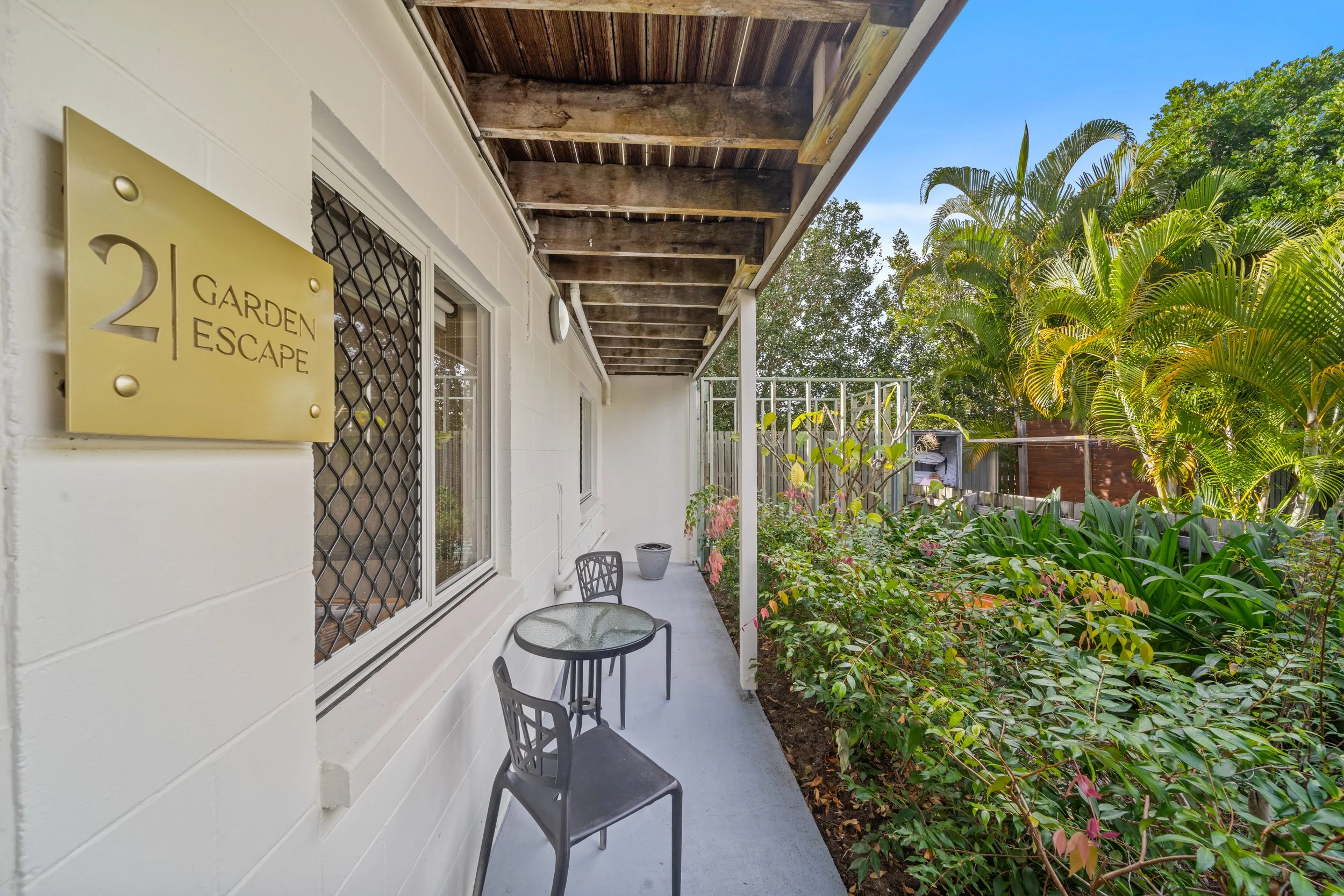 Apartment patio with two chairs, a small table, white exterior wall with windows, garden with bushes and trees, and a yellow sign reading '2 Garden Escape'.