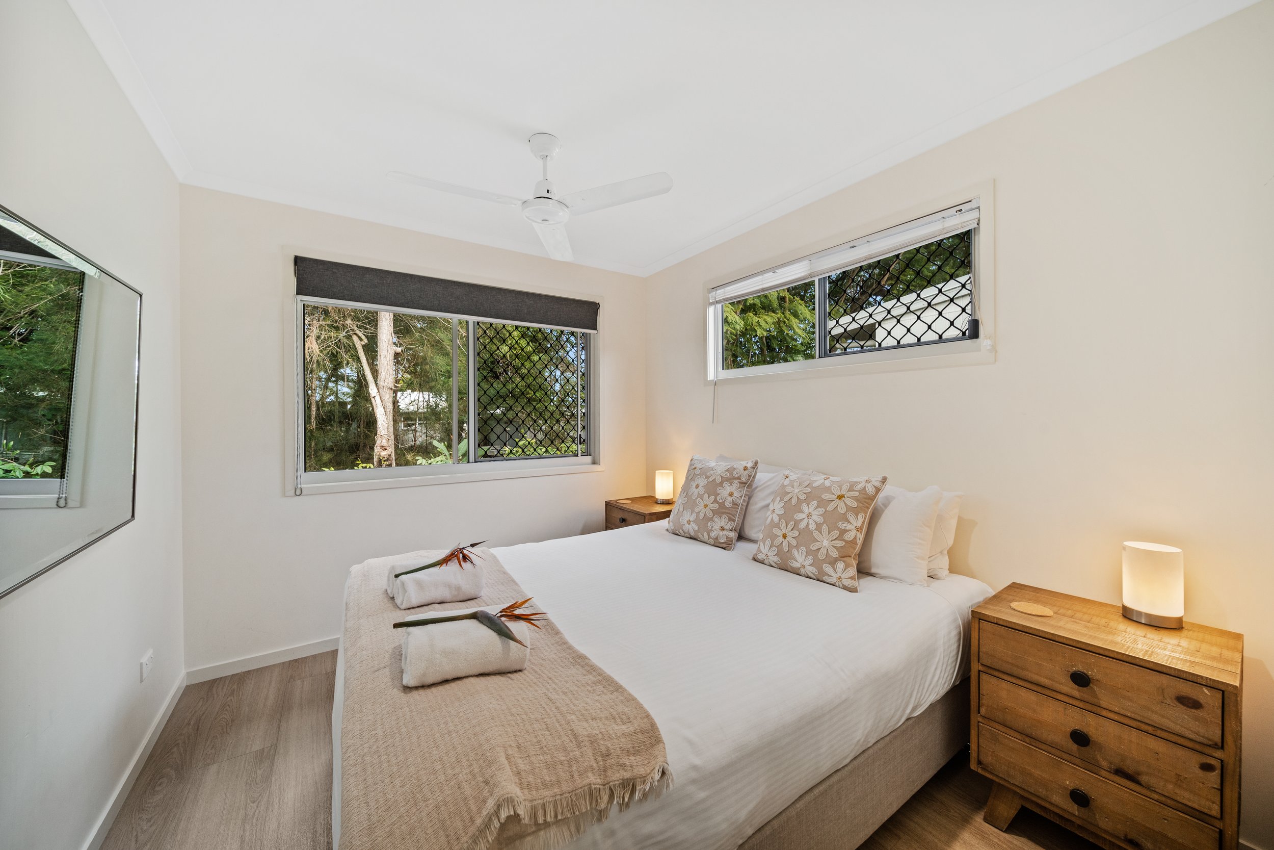 A bedroom with a white bed, beige blanket, floral pillows, a wooden nightstand with a lamp, two towels with tropical leaves, and windows with trees outside.