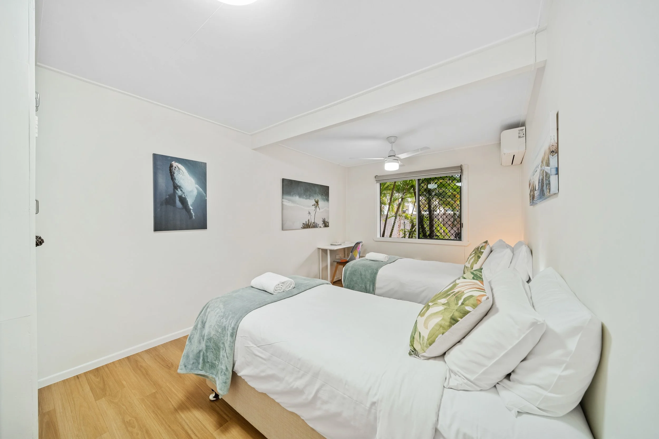 A bright hotel room with two white beds decorated with green and white pillows, a window with a view of palm trees, and wall art of ocean scenes.