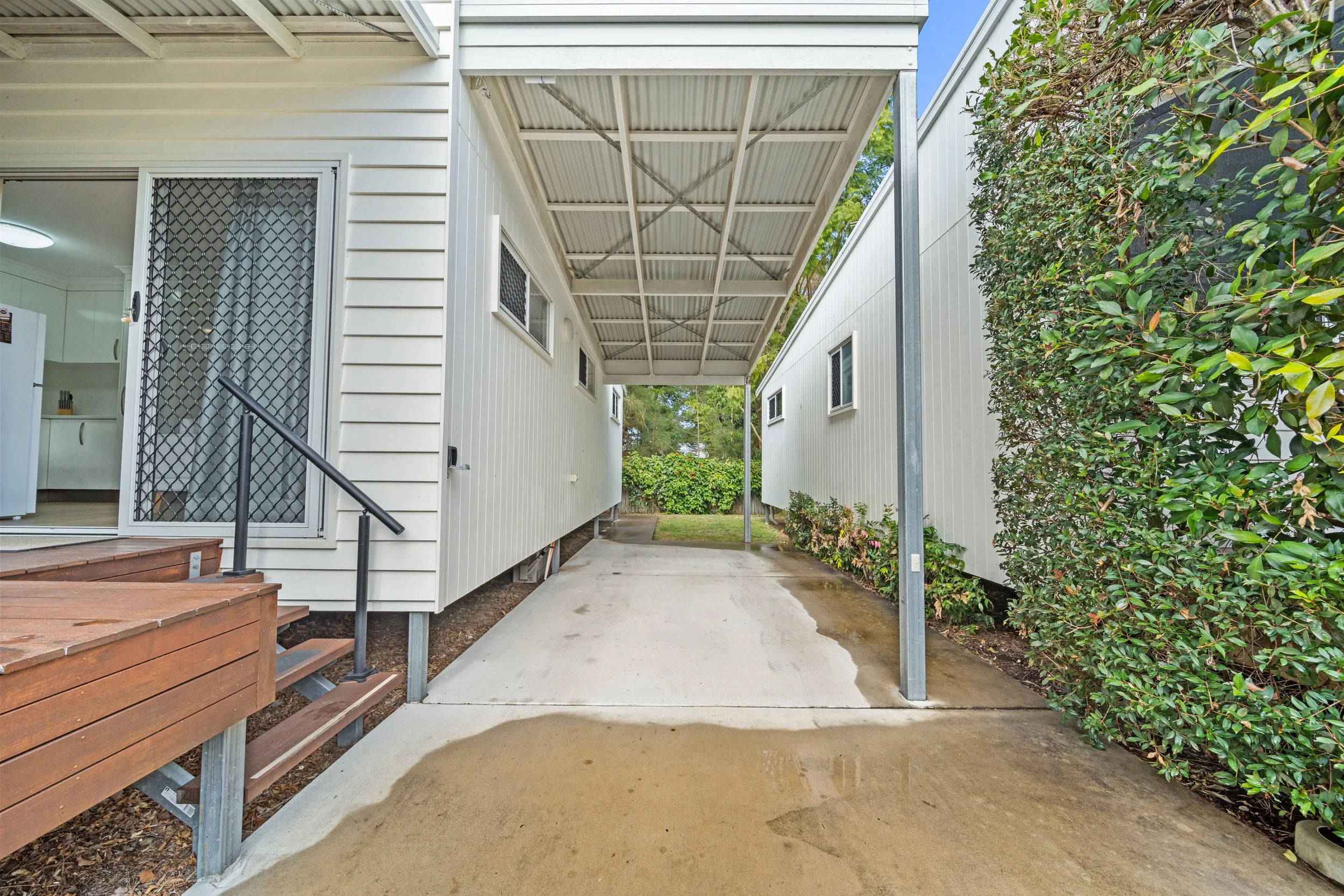 View of the outdoor area between two white modular buildings, featuring a concrete pathway, a small wooden deck, a leafy green shrub on the right, and a covered bike parking area overhead. The inside of the left building is visible through a sliding 