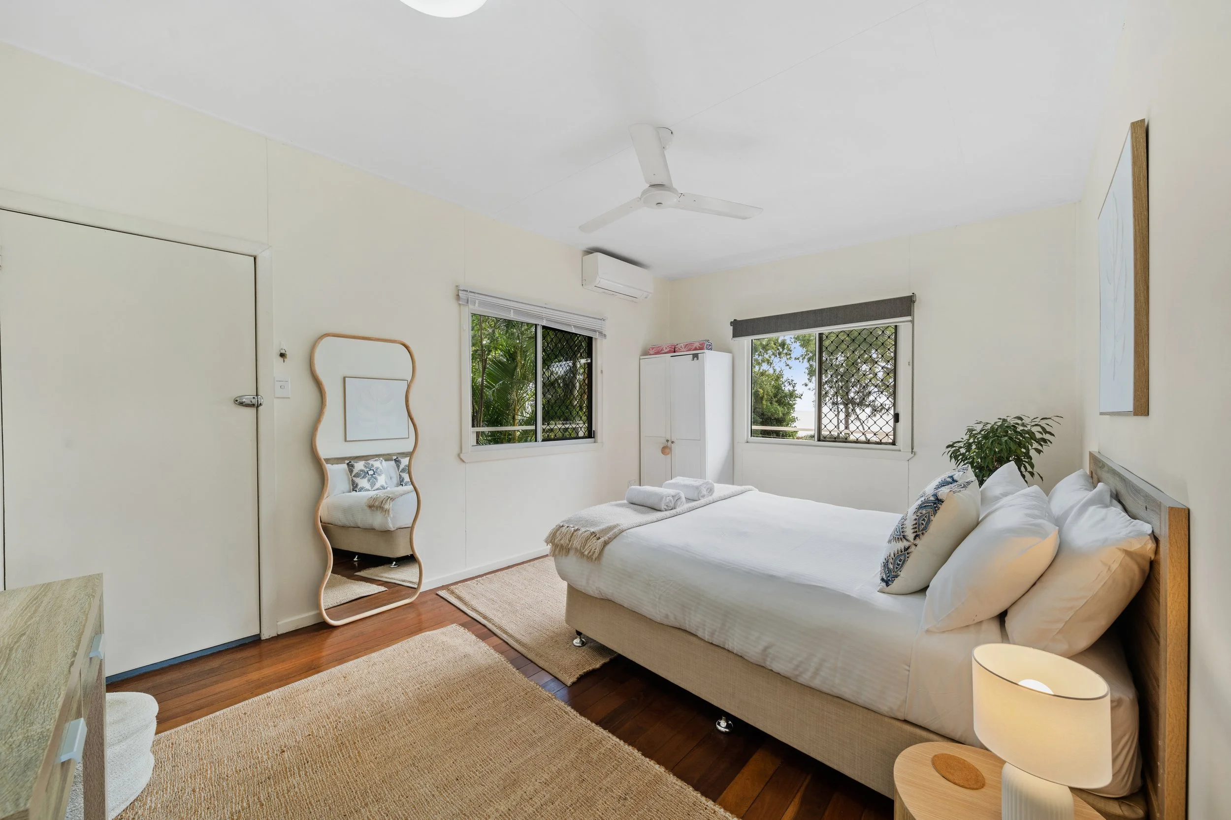 A cozy bedroom with a large bed, white linens, decorative pillows, and a wooden headboard. There are two windows with screens and blinds, a white wardrobe, a mirror with a wooden frame, a potted plant, a small side table with a lamp, and a ceiling fa
