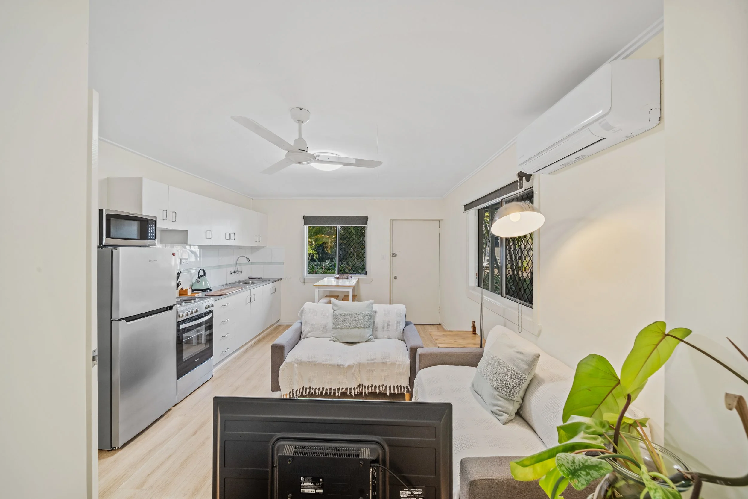 View of a living room and kitchen area with a ceiling fan, air conditioning unit, window, and a sofa with cushions.