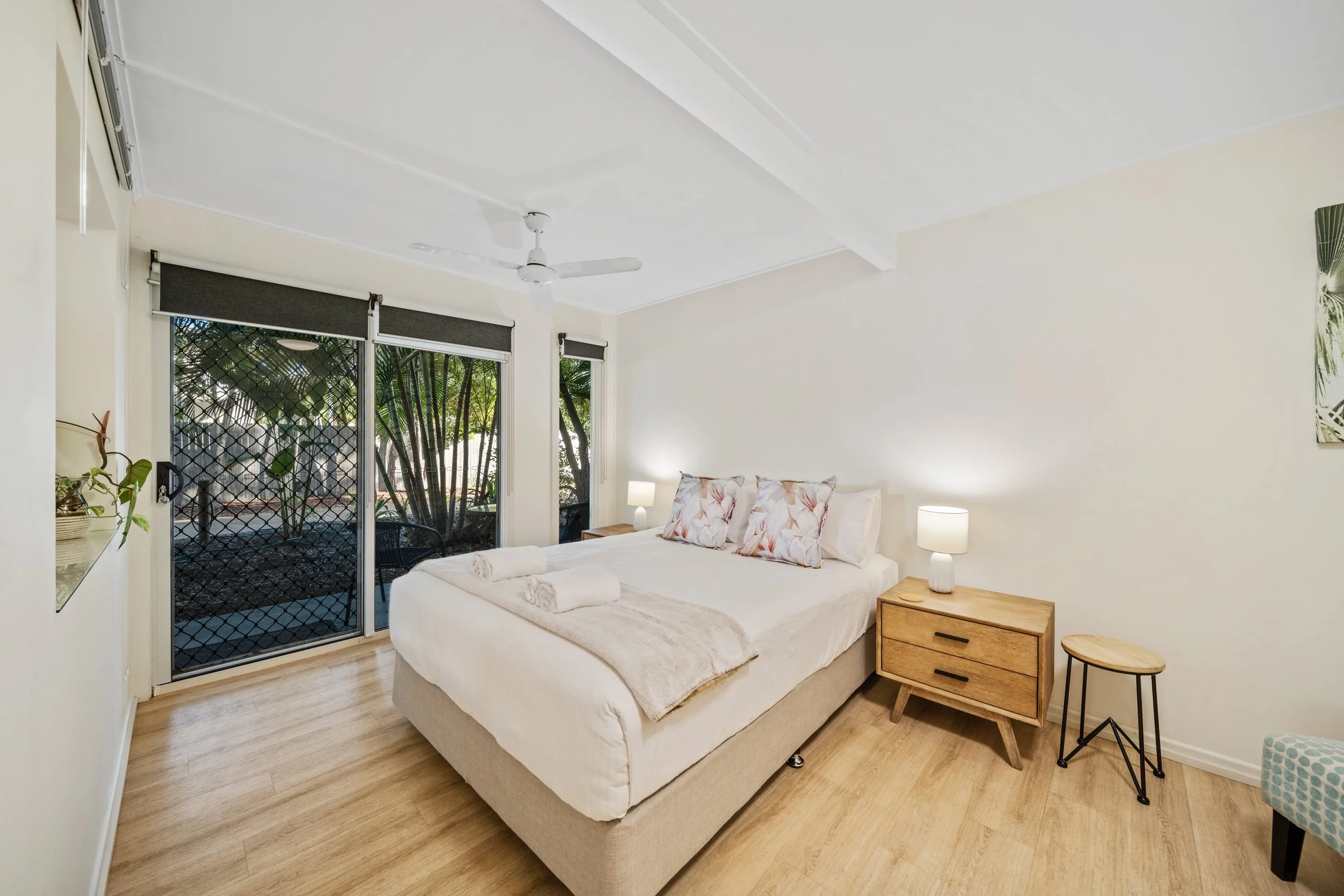 A bedroom with a bed, nightstands, lamps, and large sliding glass doors leading to an outdoor patio with trees.