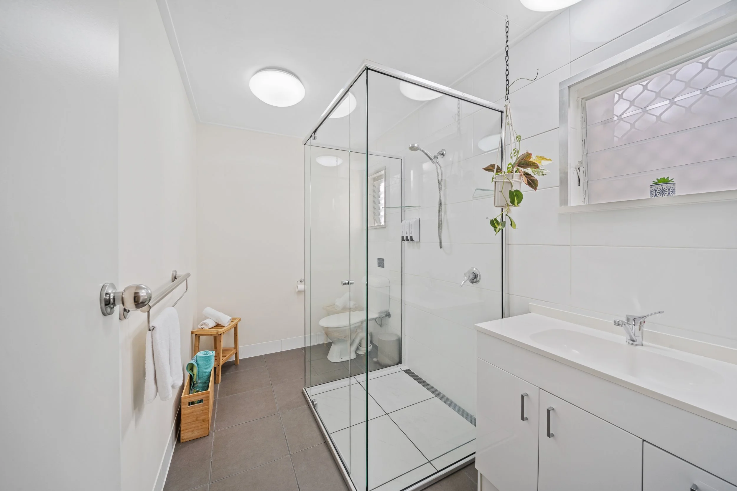 A modern bathroom with a glass shower enclosure, white walls, a white vanity with a silver faucet, and a window with a plant in a pot. There are towels hanging on a silver towel bar and a small wooden stool with rolled towels. The ceiling has built-i