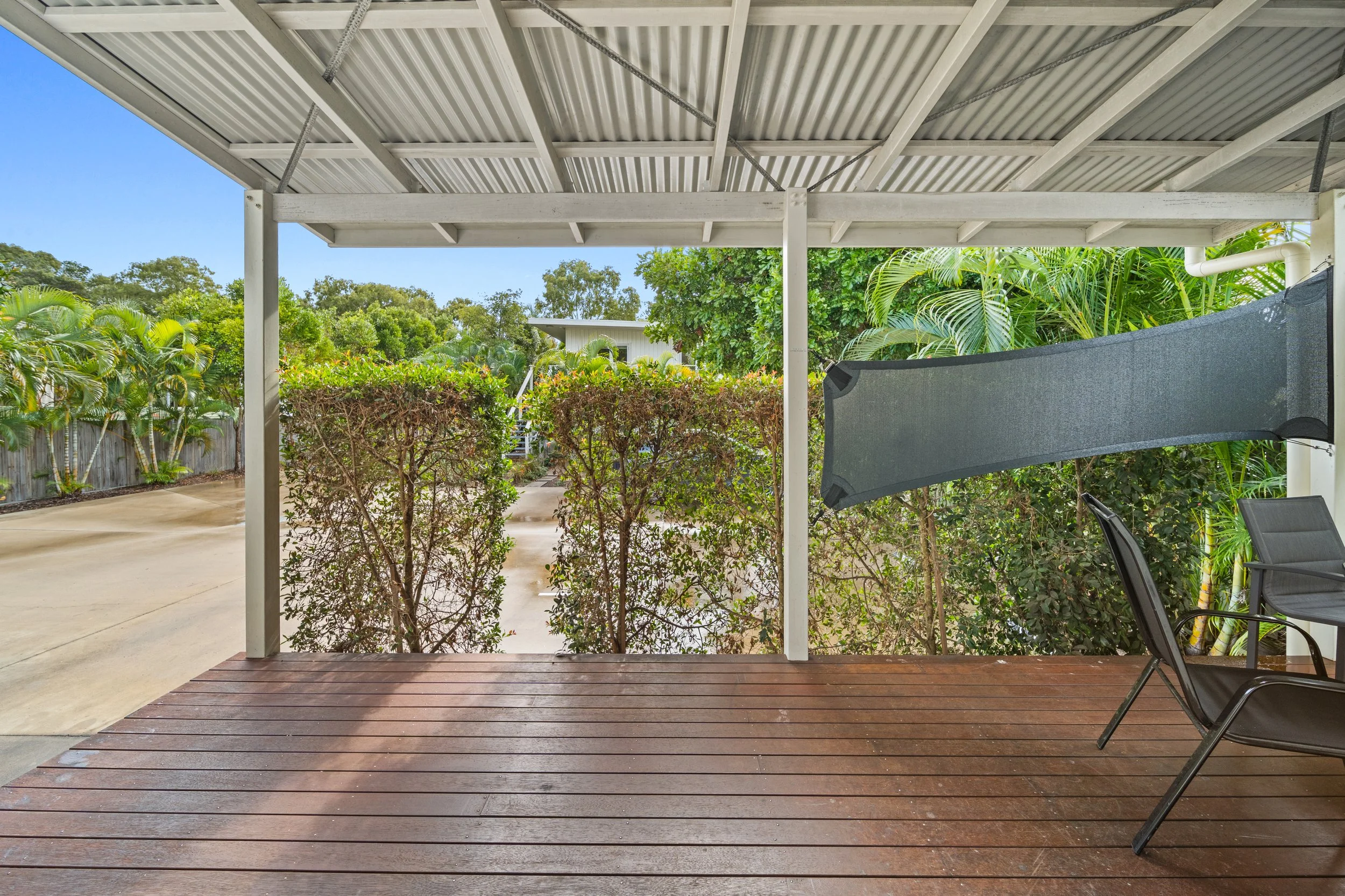 A covered backyard patio with a wooden deck, black chairs, and a shade screen, surrounded by lush green bushes and trees under a blue sky.