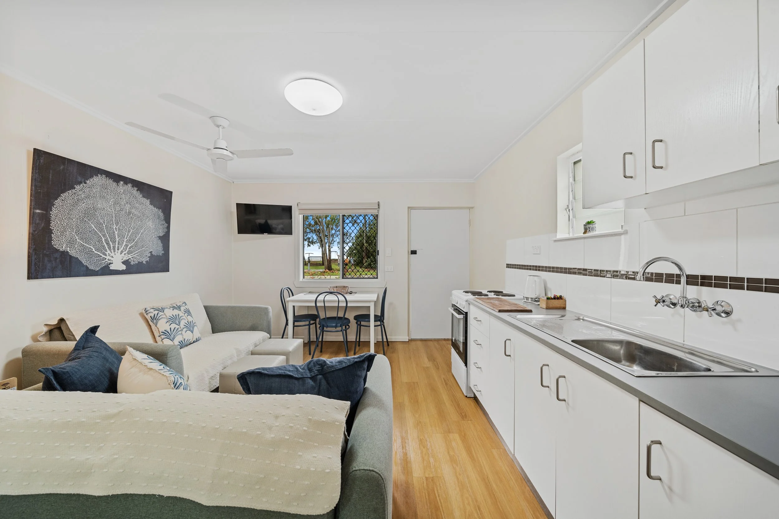 A living room and kitchen area with white walls and ceiling, wooden floor, a white sofa with patterned cushions, a wall-mounted TV, a small dining table with three chairs, a window with a view of trees, and white kitchen cabinets with silver handles 