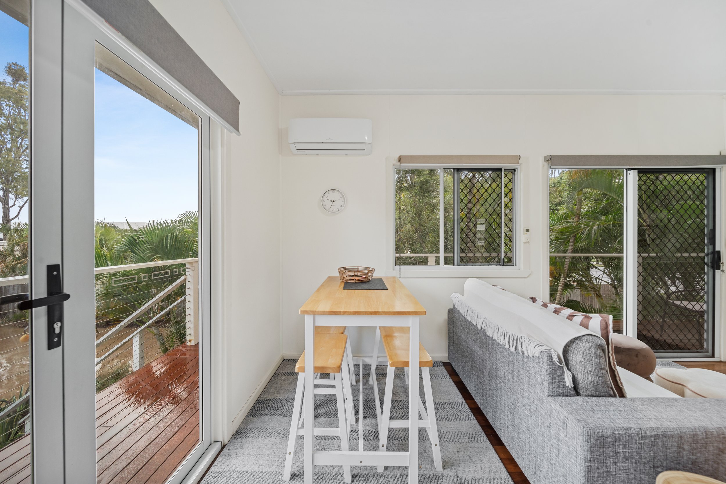 A bright living space with a sliding glass door leading to a wooden porch with trees outside. Inside, there is a gray sofa with a striped throw, a wooden dining table with matching benches, a wall clock, and an air conditioning unit. The room has whi