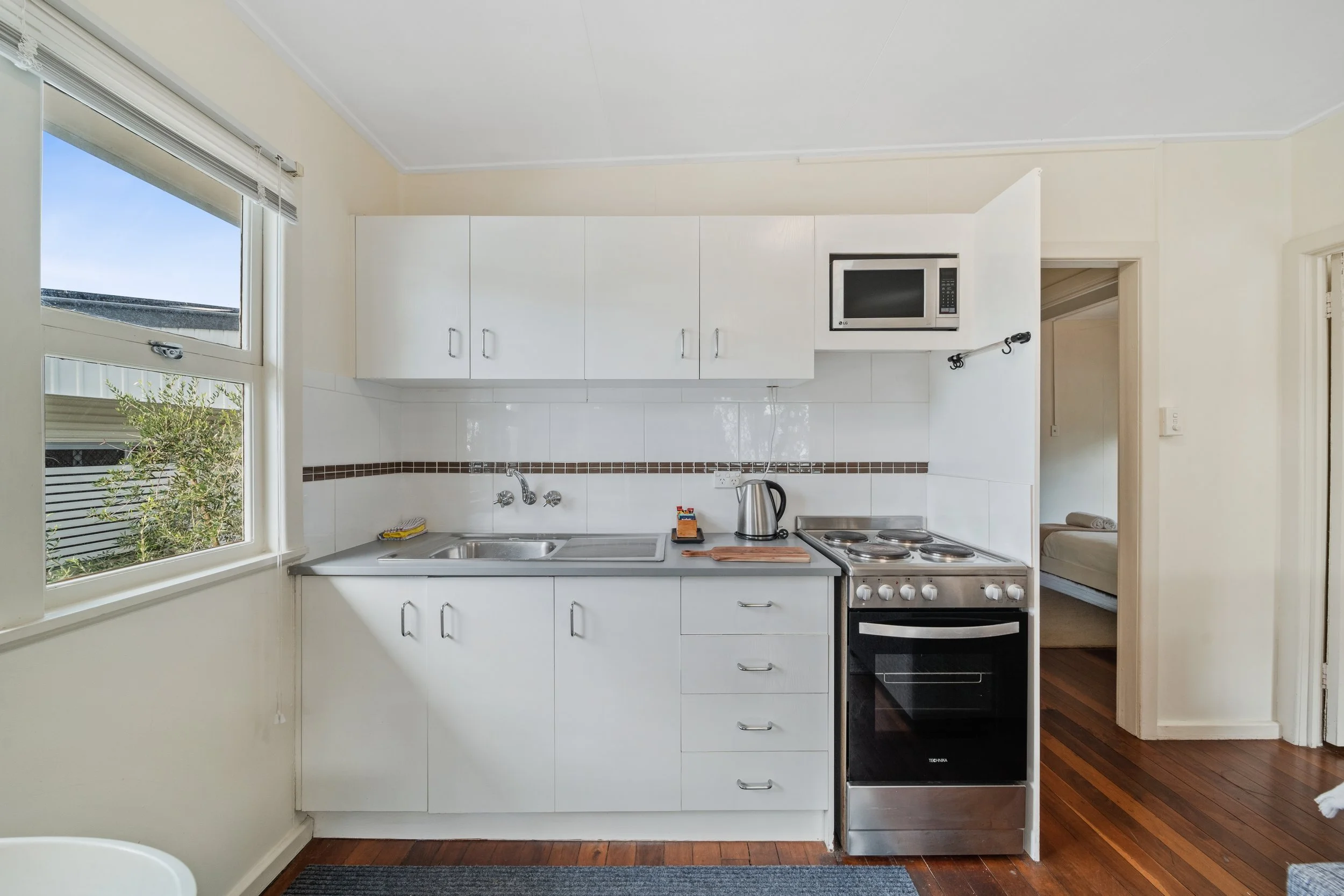 A small kitchen with white cabinets, a window, and a stove. Contains a kettle, a cutting board, a microwave, and a washing area with a sink. Adjacent room has a bed.