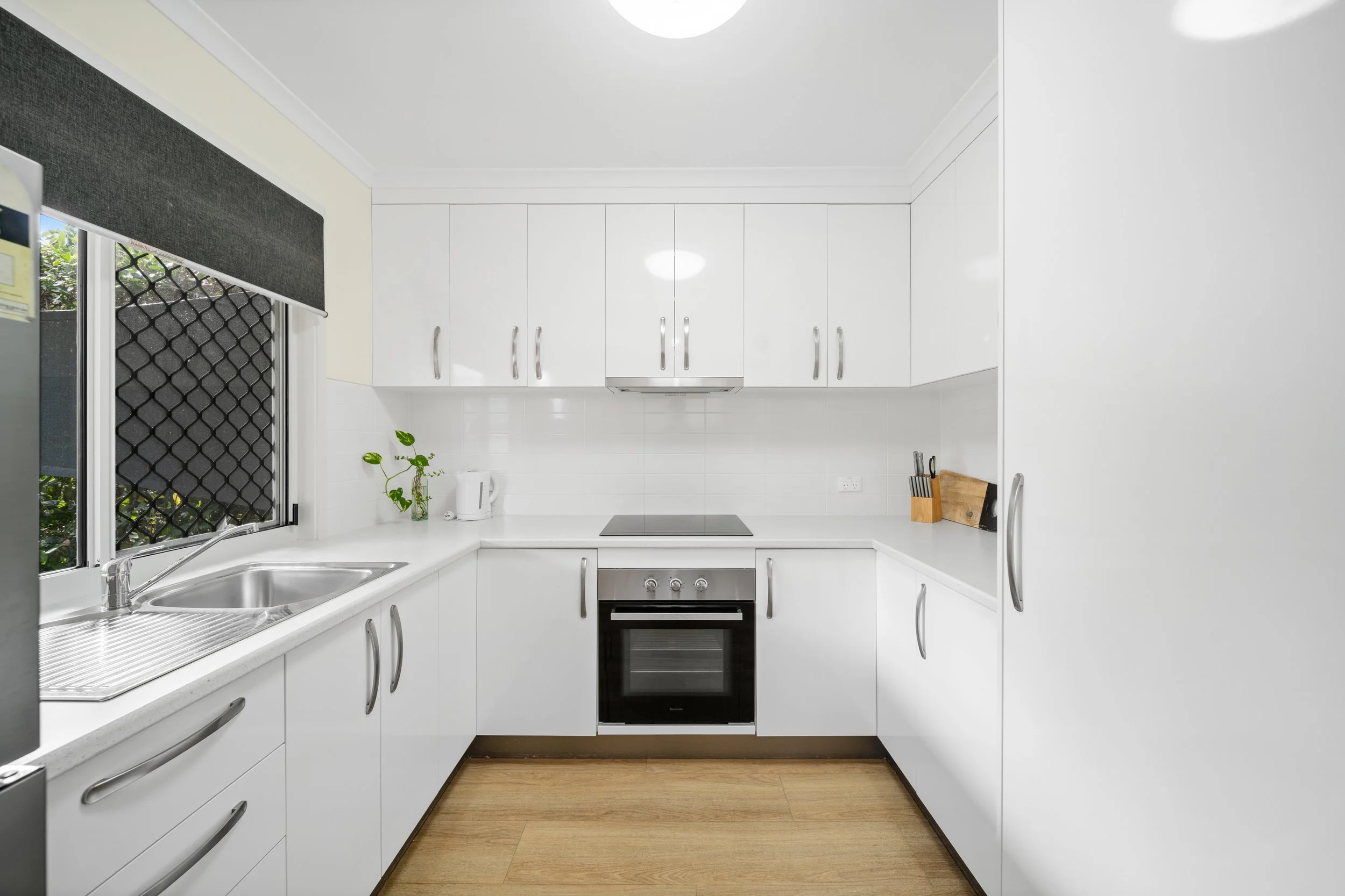 A modern kitchen with white cabinets, a stainless steel oven, a black cooktop, a white countertop, a sink next to a window with a black security screen, a white kettle, a small plant, and a set of knives on a wooden block.