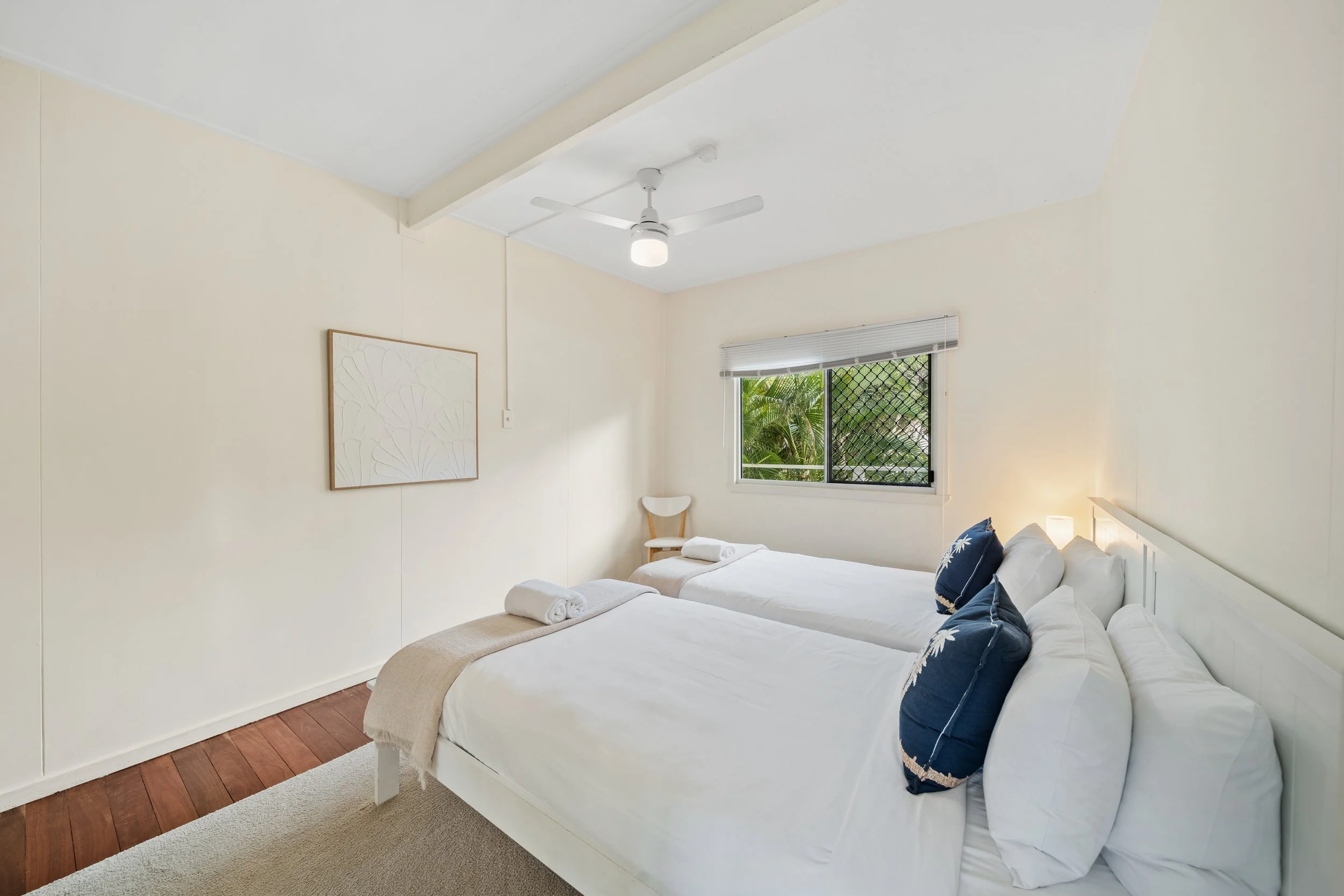 A bedroom with two single beds covered in white linens, decorated with navy blue pillows with white floral patterns. There is a window with a security screen and white blinds, and a ceiling fan with a light. The walls are painted a light color, and a
