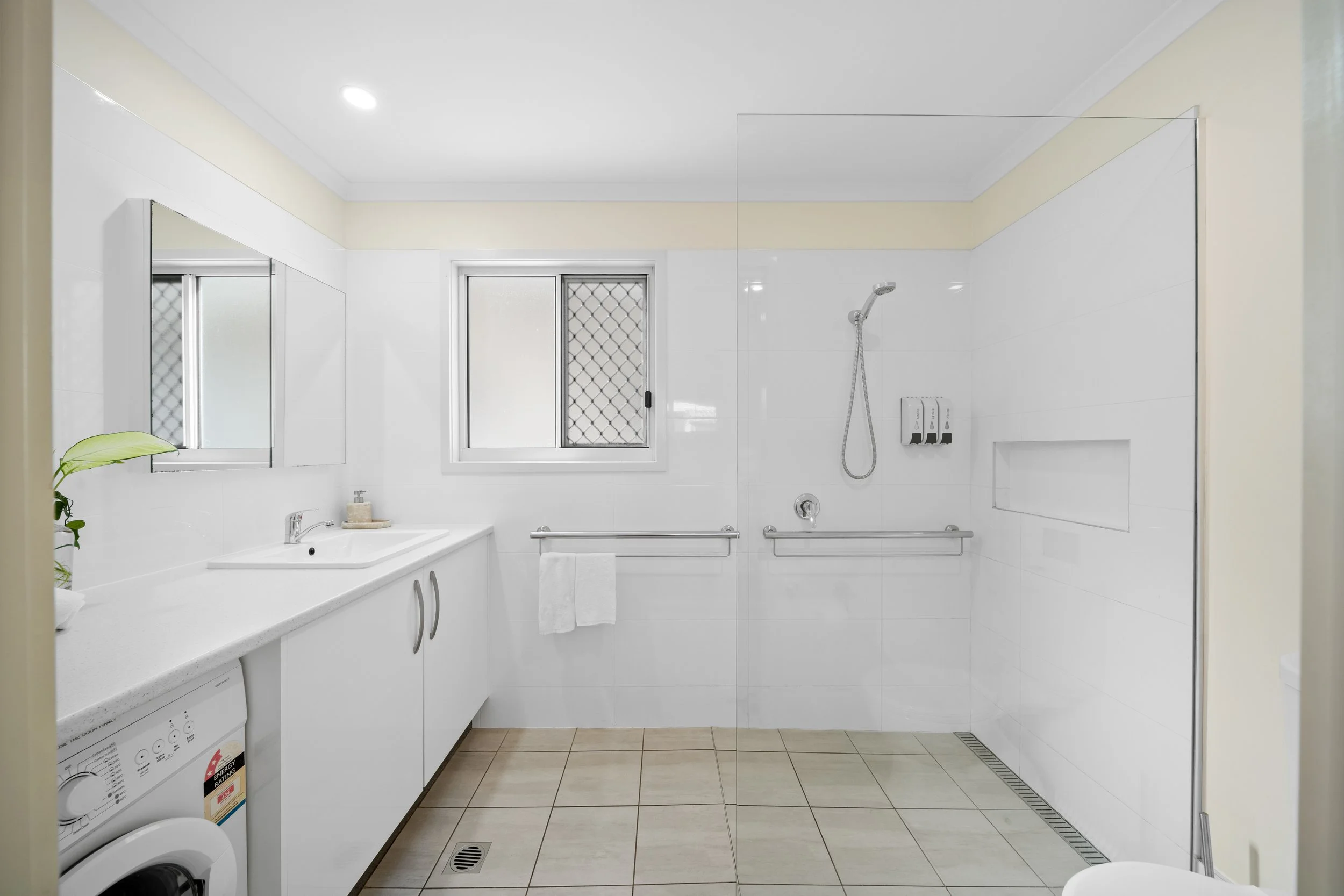 A bright, white bathroom with a shower area that has frosted windows, a rain shower head, and grab bars. There is a vanity with a countertop, faucet, and a mirror above it. The bathroom also contains a washing machine and beige floor tiles.