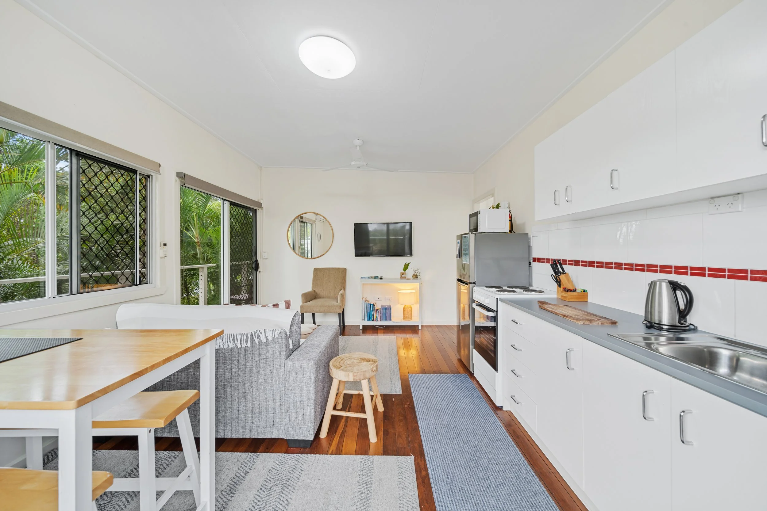 A bright, modern living space with large windows overlooking greenery, a beige chair, a flat-screen TV on the wall, and a kitchen area with white cabinets, a stove, microwave, and small appliances.