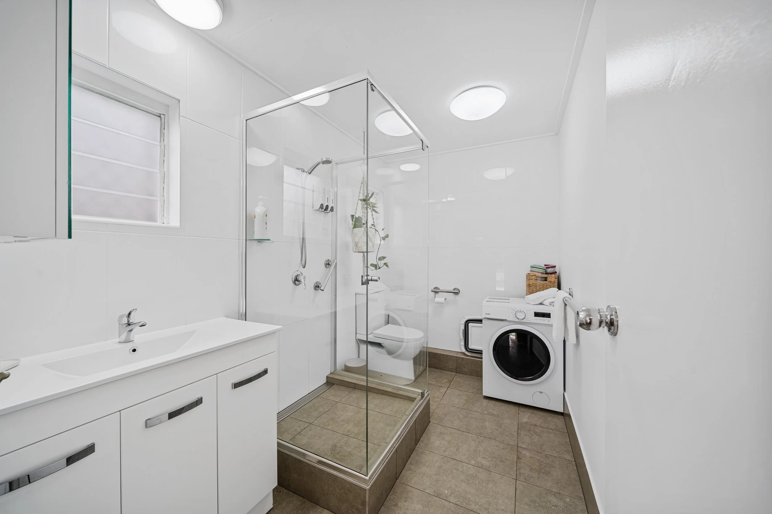 A modern bathroom with a glass-enclosed shower, white vanity with two sinks, a window, a washing machine, a wicker basket, and various toiletries and towels. The room has white walls and ceiling with circular ceiling lights.