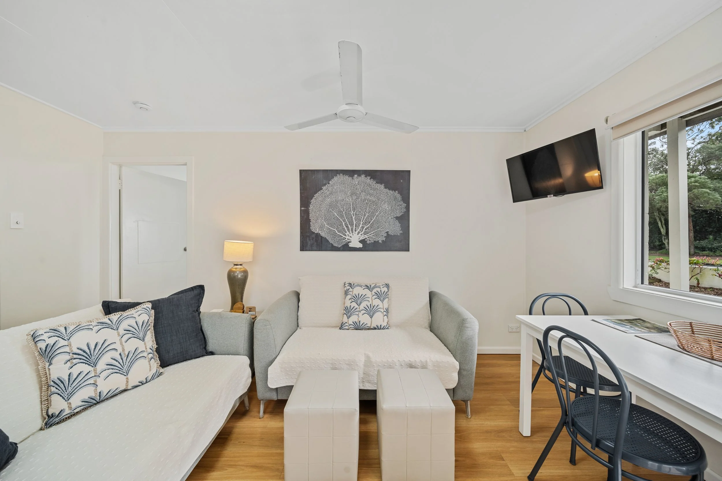 Living room with light-colored walls, a white ceiling fan, a wall art of a tree, a table lamp, white sofa with decorative pillows, a small white table, a TV mounted on the wall, and a sliding door with a window showing trees outside.