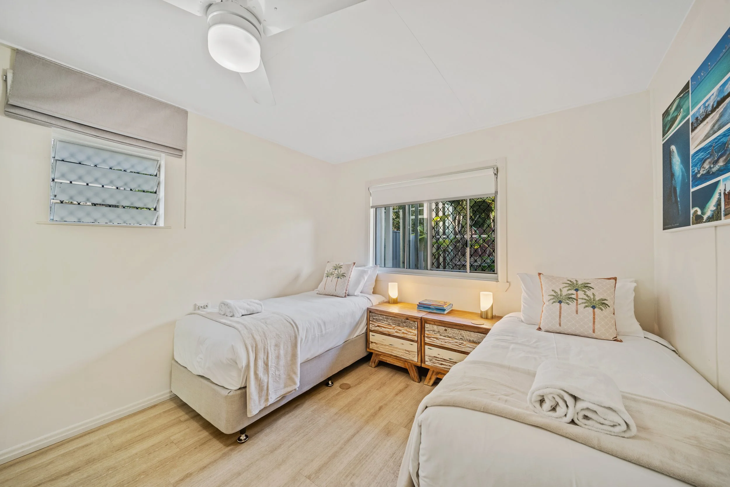 A bright bedroom with two single beds, each with white bedding and rolled towels. There's a wooden nightstand with a lamp and books between the beds. The room has a window with white blinds and a second window with a security grille, decorated with o
