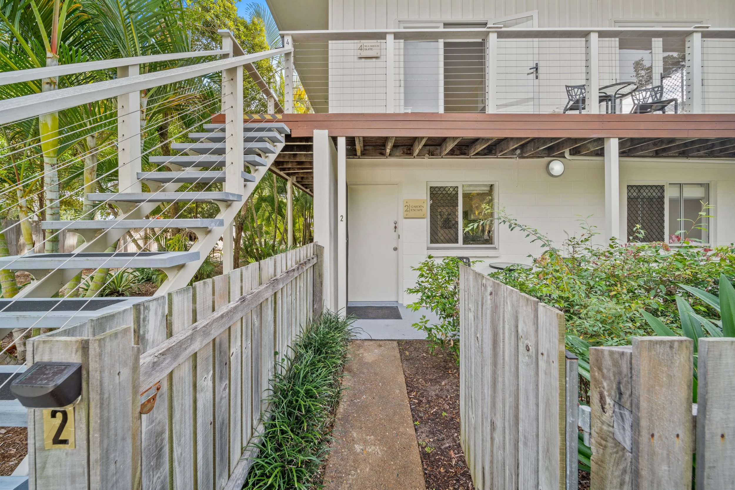 Front walkway leading to a two-story apartment with stairs on the left side, surrounded by garden with wooden fencing and greenery.