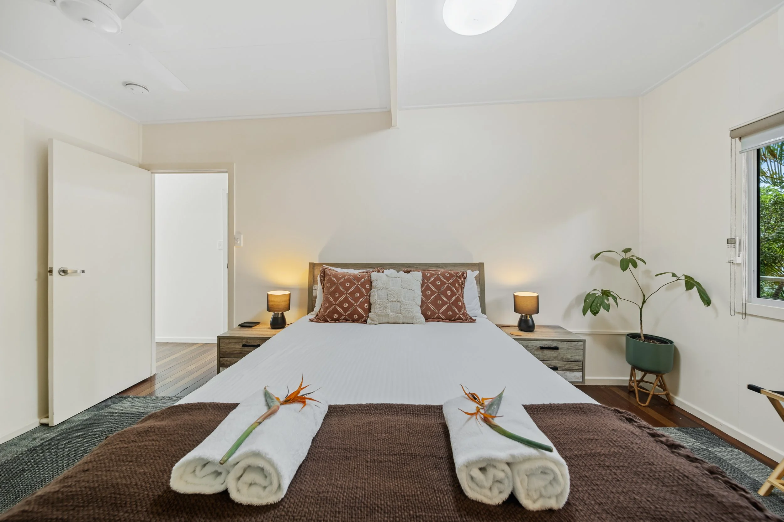 A bedroom featuring a bed with a white bedspread, decorative pillows, and towels arranged with tropical flowers. The room has bedside tables with lamps, a potted plant, and a window with blinds. There's an open door on the left and ceiling lights.