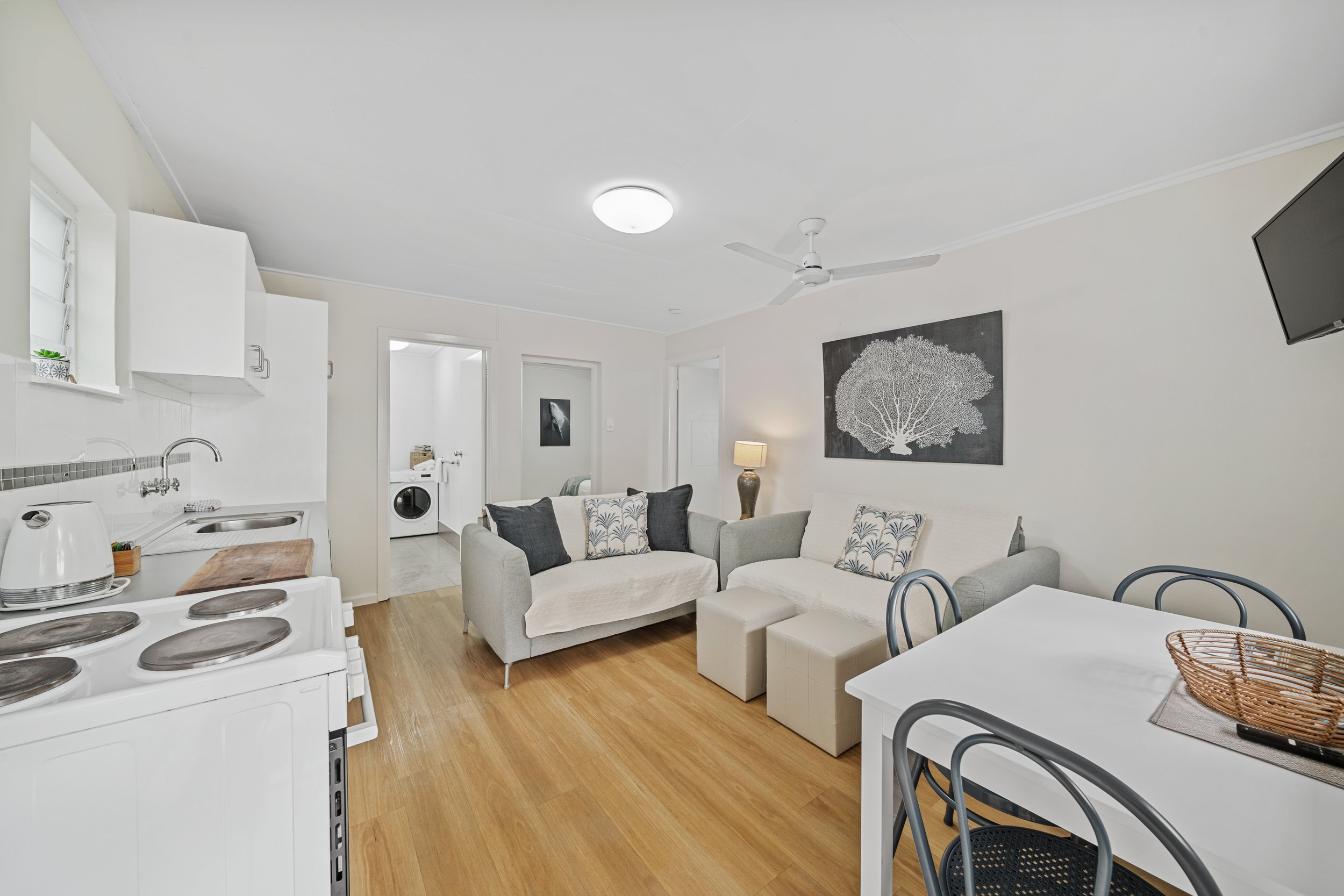 Living room with white walls, a white ceiling fan, a black and white wall art, beige sofas with patterned pillows, a lamp, a television mounted on the wall, a white kitchen counter, a stovetop, and a dining table with black chairs.