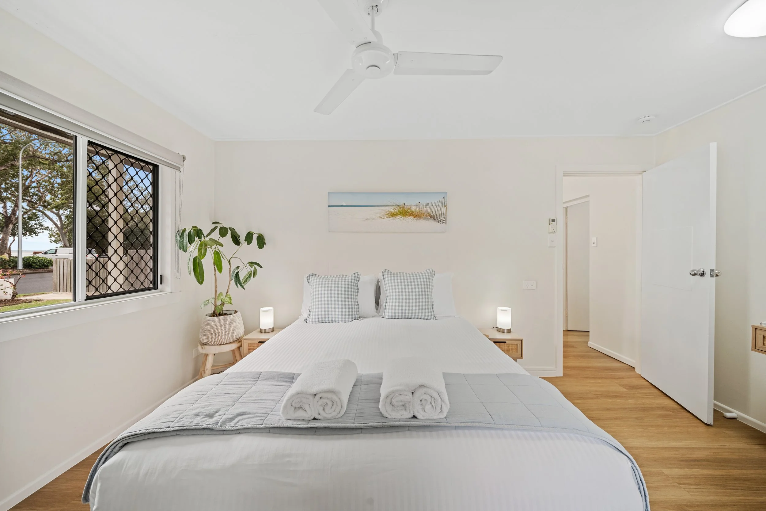 A bedroom with a bed covered in white linens and three rolled white towels on top. There are two checkered pillows on the bed. To the left, there's a small round wooden table with a potted plant. Two small lamps are on either side of the bed. The roo