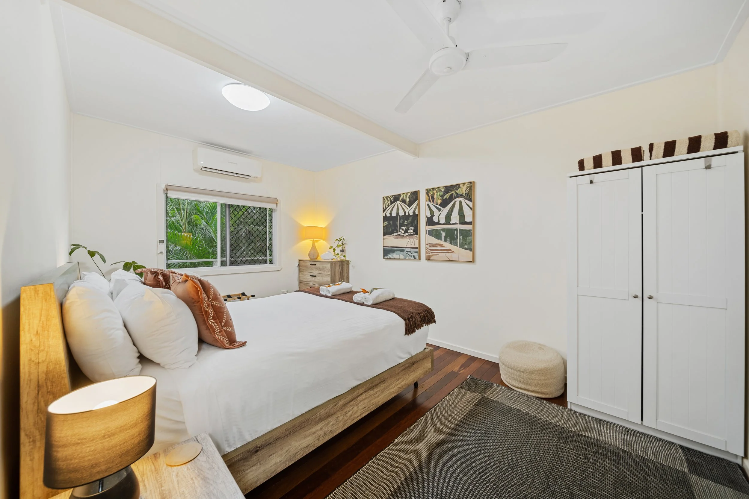 A bedroom with a wooden bed, white bedding, pillows, and chocolates. There is a window with a view of green plants, wall art, a nightstand with a lamp, a appearing air conditioning unit, and a white closet door.