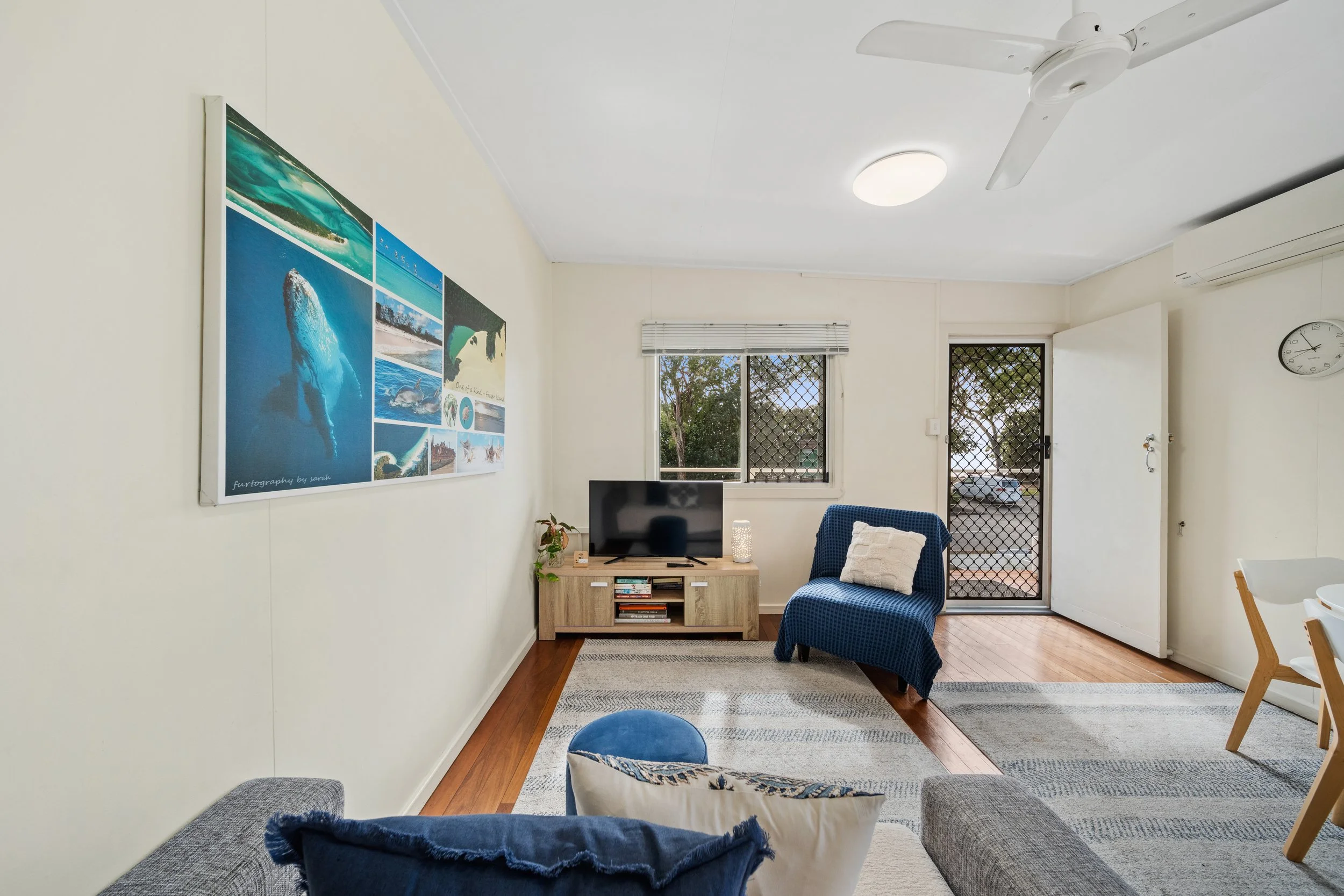 A living room with a white ceiling, ceiling fan, and round ceiling light. A large window with blinds and security screen door shows trees outside. A wall features a collage of ocean-themed photographs. Furniture includes a television on a wooden stan
