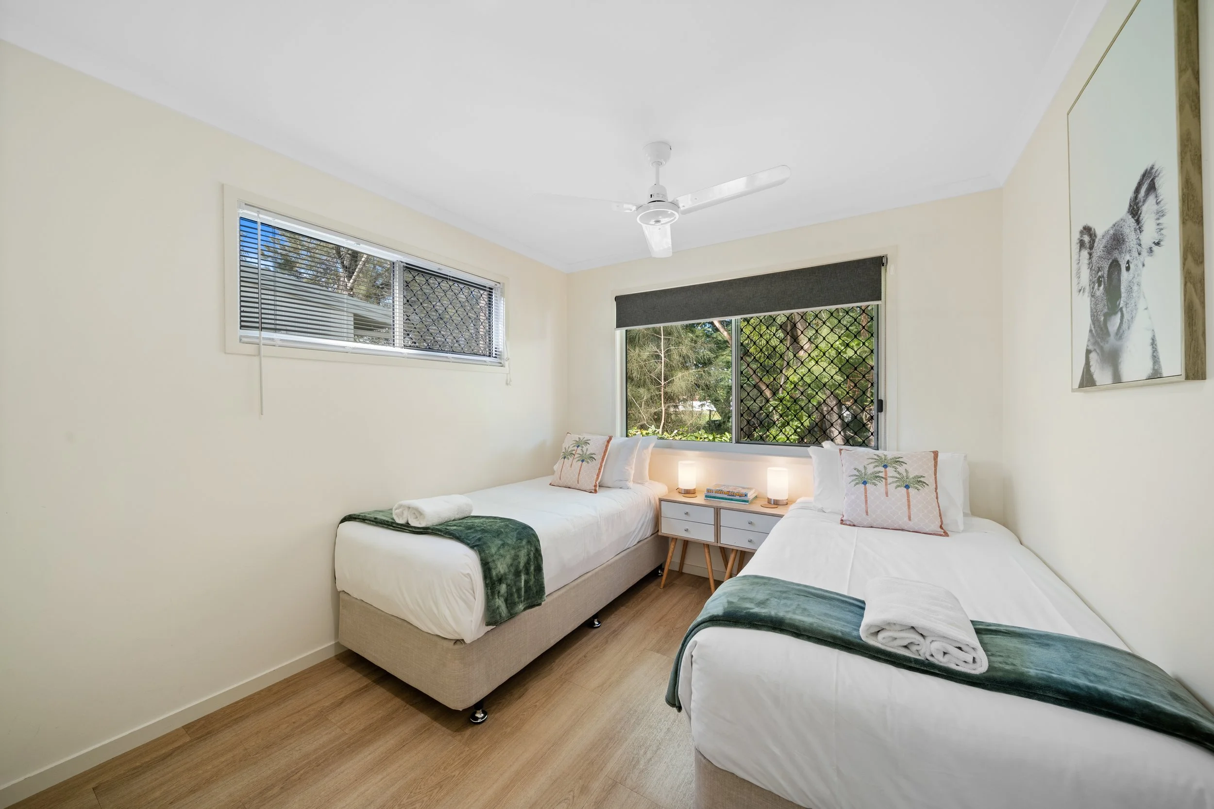 A bedroom with two beds parallel to each other, each with white linens, green throws, and rolled towels, a nightstand with lamps and books, windows with blinds and a view of trees, and a white ceiling fan.