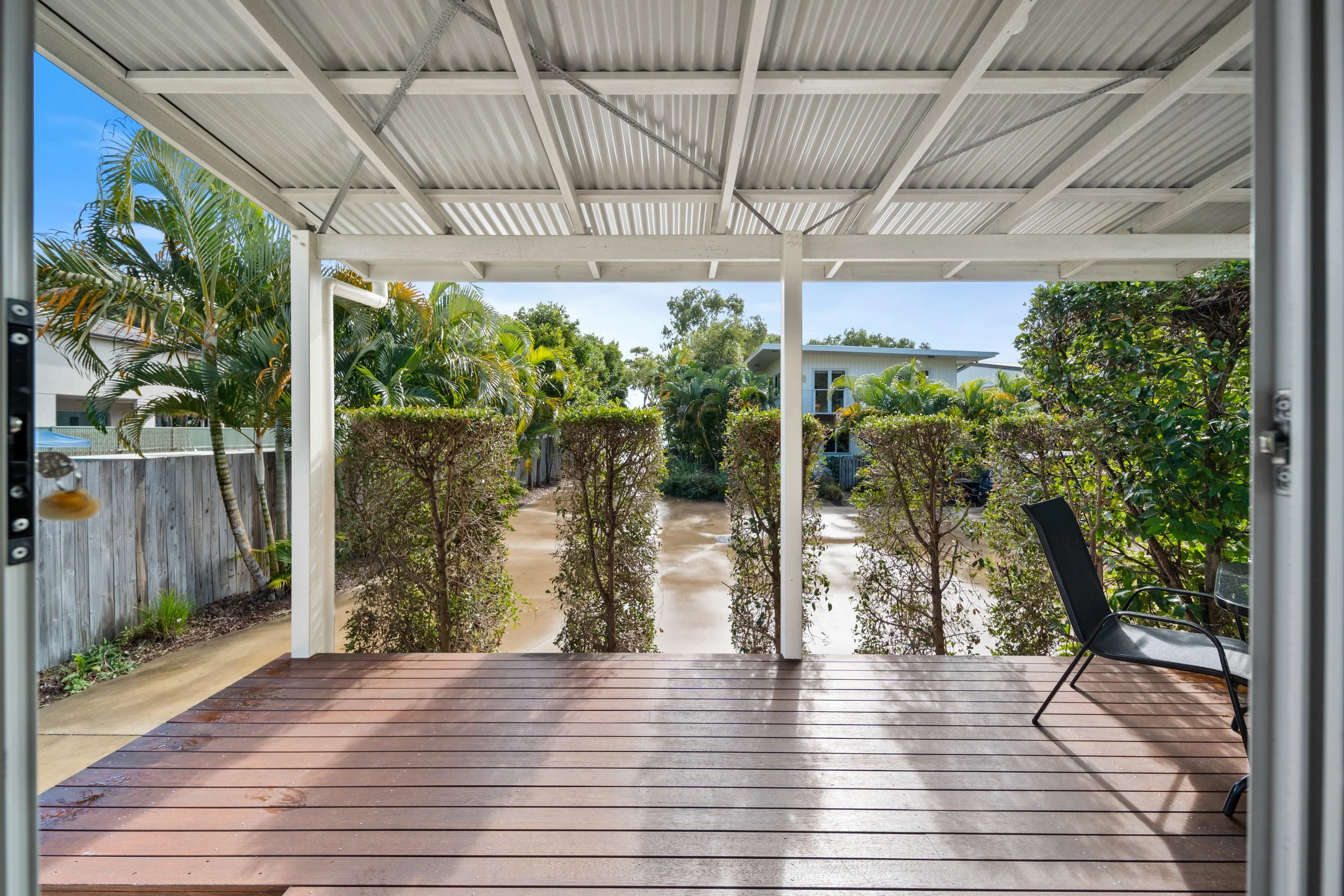 View of a backyard patio with a privacy fence, lush green bushes, and trees, viewed from under a roofed porch with a wooden deck, two black chairs, and a garden hose.